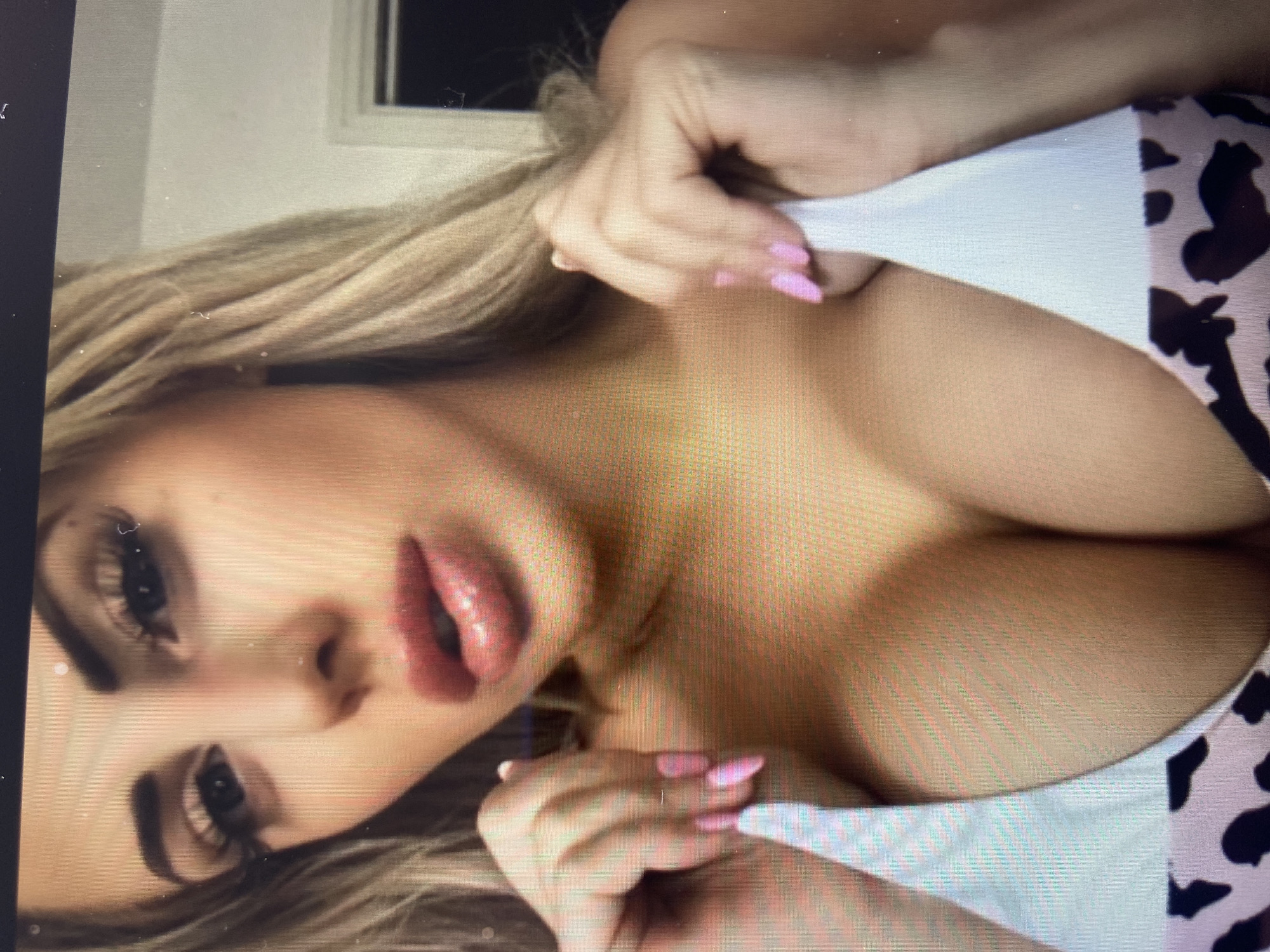 A close-up, head-and-shoulders shot shows a woman with blonde hair and light skin. She is wearing a white and pink leopard-print bikini top. Her lips are painted with a shimmery pink lipstick, and she has long, dark eyelashes. Her fingernails are painted bright pink. She is holding the straps of her bikini top with both hands. The background is out of focus, but appears to be a room with a white door frame on the right. The image is pixelated, suggesting it was taken from a screen.