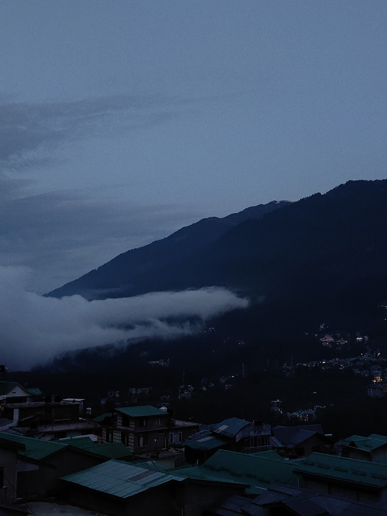 The image depicts a mountain village scene at dusk, likely Vashisht, India. The foreground features a collection of houses with distinctive green roofs, indicating a residential area. The architecture appears to be traditional, with multiple stories and varied rooflines. The houses are clustered together, suggesting a dense settlement.
Beyond the village, the mid-ground is dominated by a steep, dark mountain slope. A band of white clouds or mist is visible clinging to the lower part of the mountain, obscuring a portion of the landscape. The mountain is sparsely dotted with trees, forming a darker silhouette against the slightly lighter sky.
The background shows a vast, dark mountain range that stretches across the horizon. The sky above is a deep, muted blue, with some lighter clouds present, indicating the twilight hour. Tiny lights from the village are visible scattered on the hillside, hinting at a larger settlement or town further up the mountain. The overall atmosphere is serene and cool, characteristic of an evening in a mountainous region. No people are visible in the image.