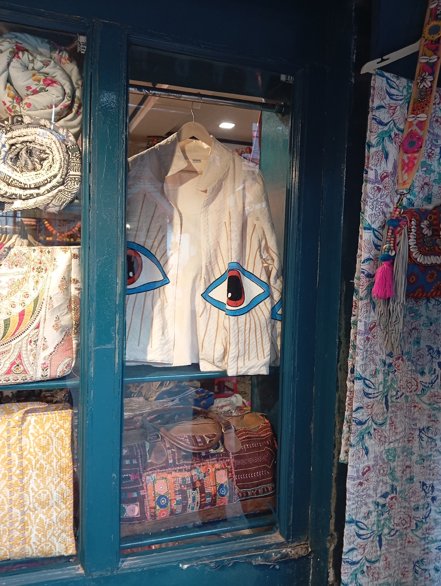 A vibrant shop window in the Unknown city showcases a diverse collection of textiles and fashion items. The window's blue frame divides the display into distinct sections. On the left, stacked quilted materials in various floral and geometric patterns create a cozy, layered effect. The center section features a light-colored jacket hanging on a hanger, adorned with stylized eye designs, set against a backdrop of more textiles. Below the jacket, patterned bags add to the textural richness of the display. To the right, a long piece of floral fabric hangs alongside a decorative bag with tassels and embellishments, adding a touch of bohemian flair. The setting and diverse array of textiles suggest a bohemian or artisan boutique, appealing to customers with eclectic tastes.