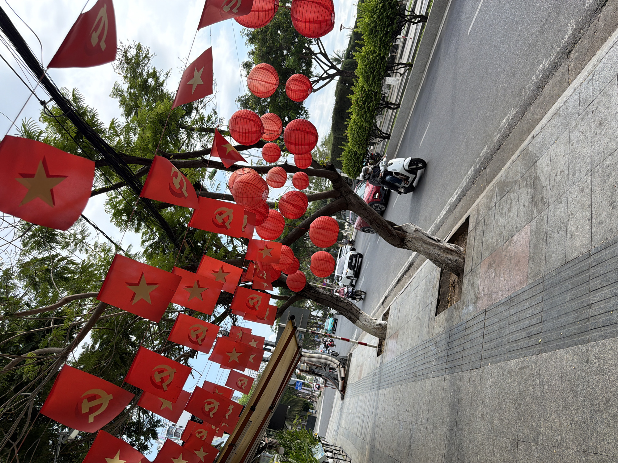 The image shows a street scene in Nha Trang Ward, Vietnam, decorated with red flags and lanterns. The flags, some with a gold star and others with a hammer and sickle emblem, are hung from trees lining the sidewalk. Red lanterns are strung across the trees. A road runs alongside the sidewalk with scooters and vehicles driving on it. The sidewalk is tiled. The sky is overcast.