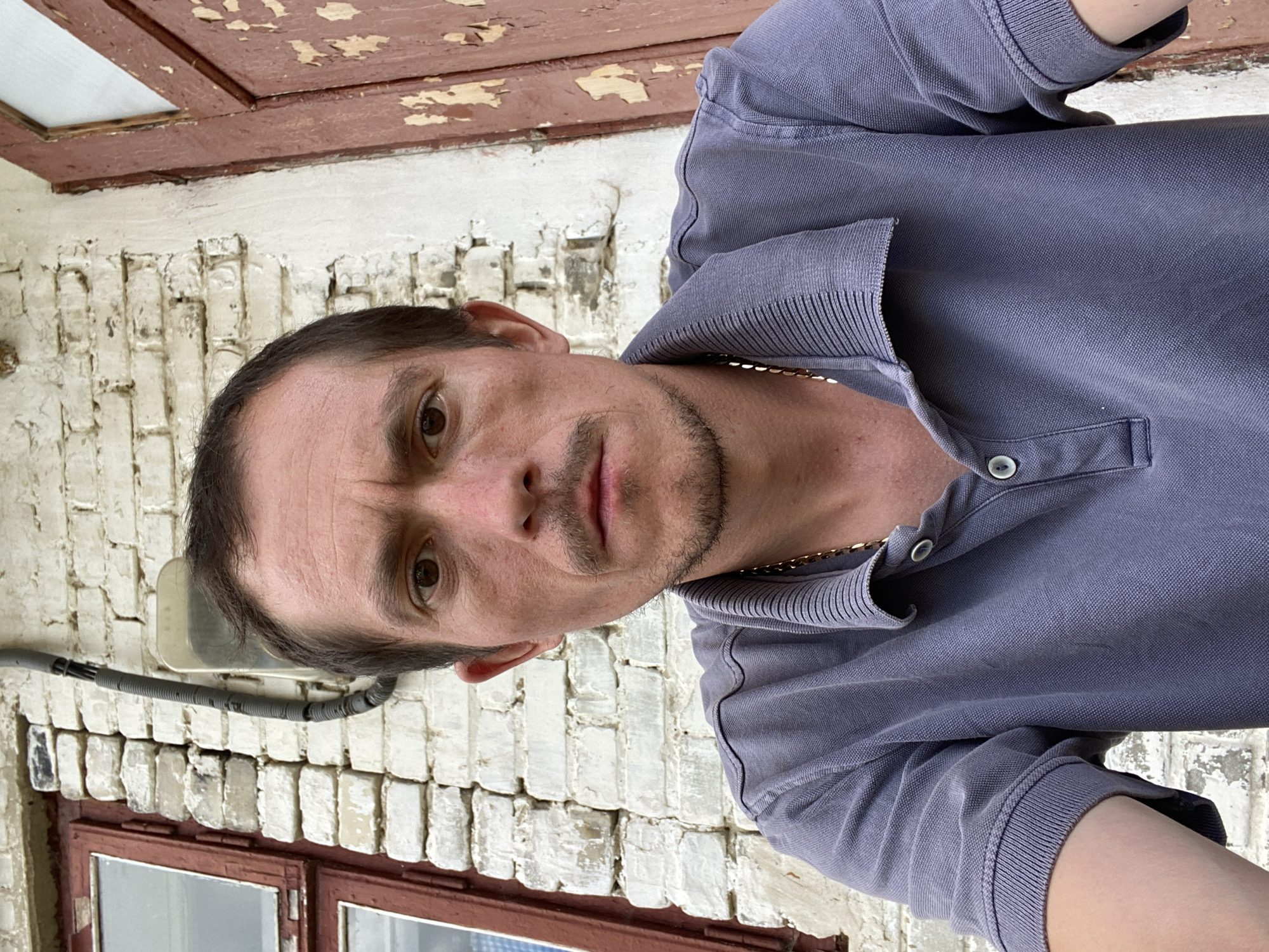 A medium shot shows a fair-skinned man with brown hair and brown eyes, wearing a dark blue polo shirt and a gold chain, looking directly at the camera. He has a thin mustache and a goatee, and his forehead is furrowed. He is positioned in front of a weathered brick wall, painted white, with visible signs of age and dirt. To the left of the man, a wooden window frame with red paint is visible. To the right, a peeling red wooden door stands adjacent to the wall. The scene appears to be outdoors, possibly in a rural or older urban setting, within the city of Mostyska, Ukraine.