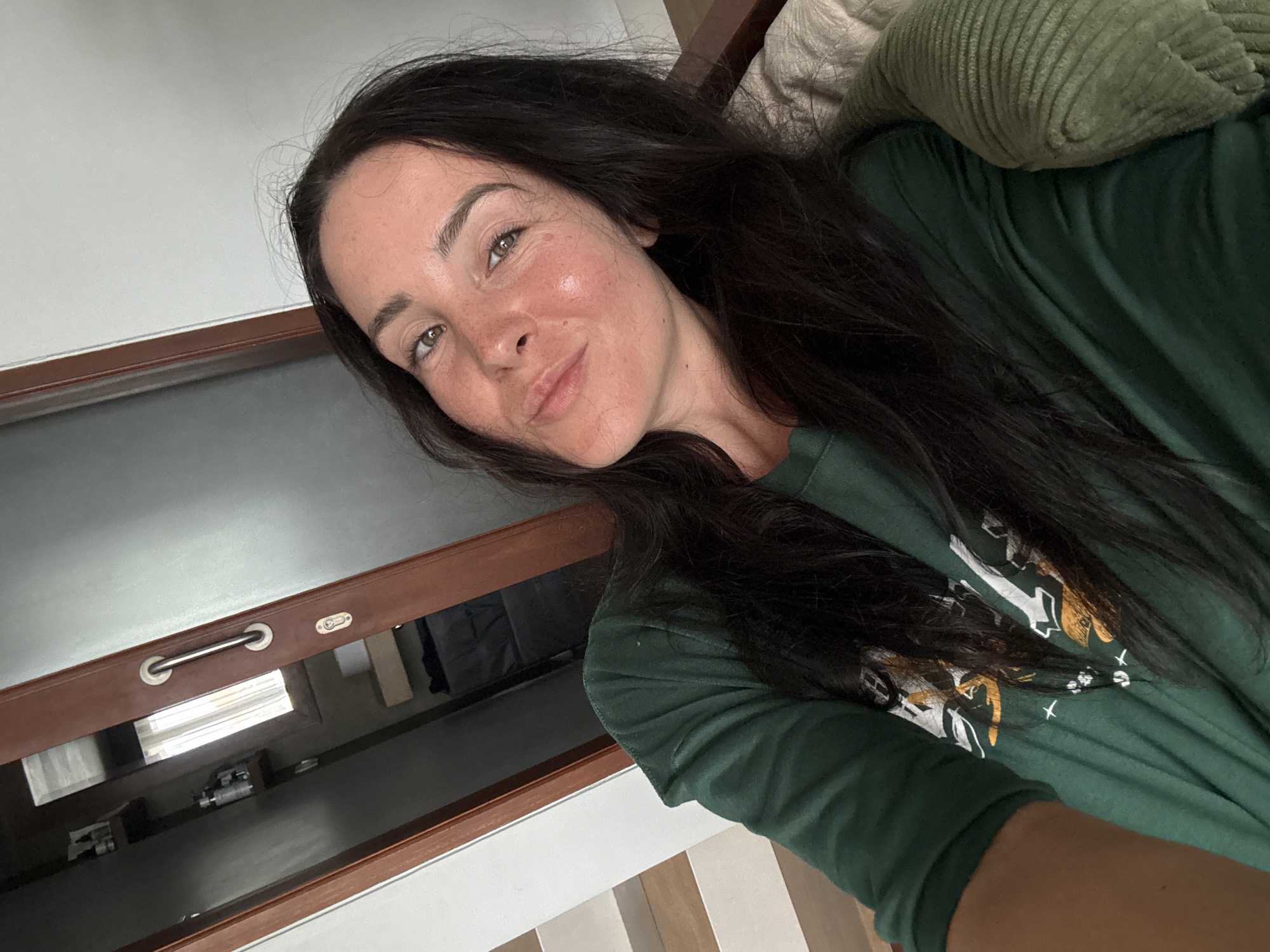 A woman with dark hair and fair skin, wearing a green t-shirt, is photographed from the chest up. She is looking at the camera with a slight smile, her eyes are green, and she has freckles. She appears to be indoors, with a wooden door frame and a staircase visible in the background. The overall lighting suggests it is daytime. There are no specific indicators to place the location in North Kuta, Indonesia.