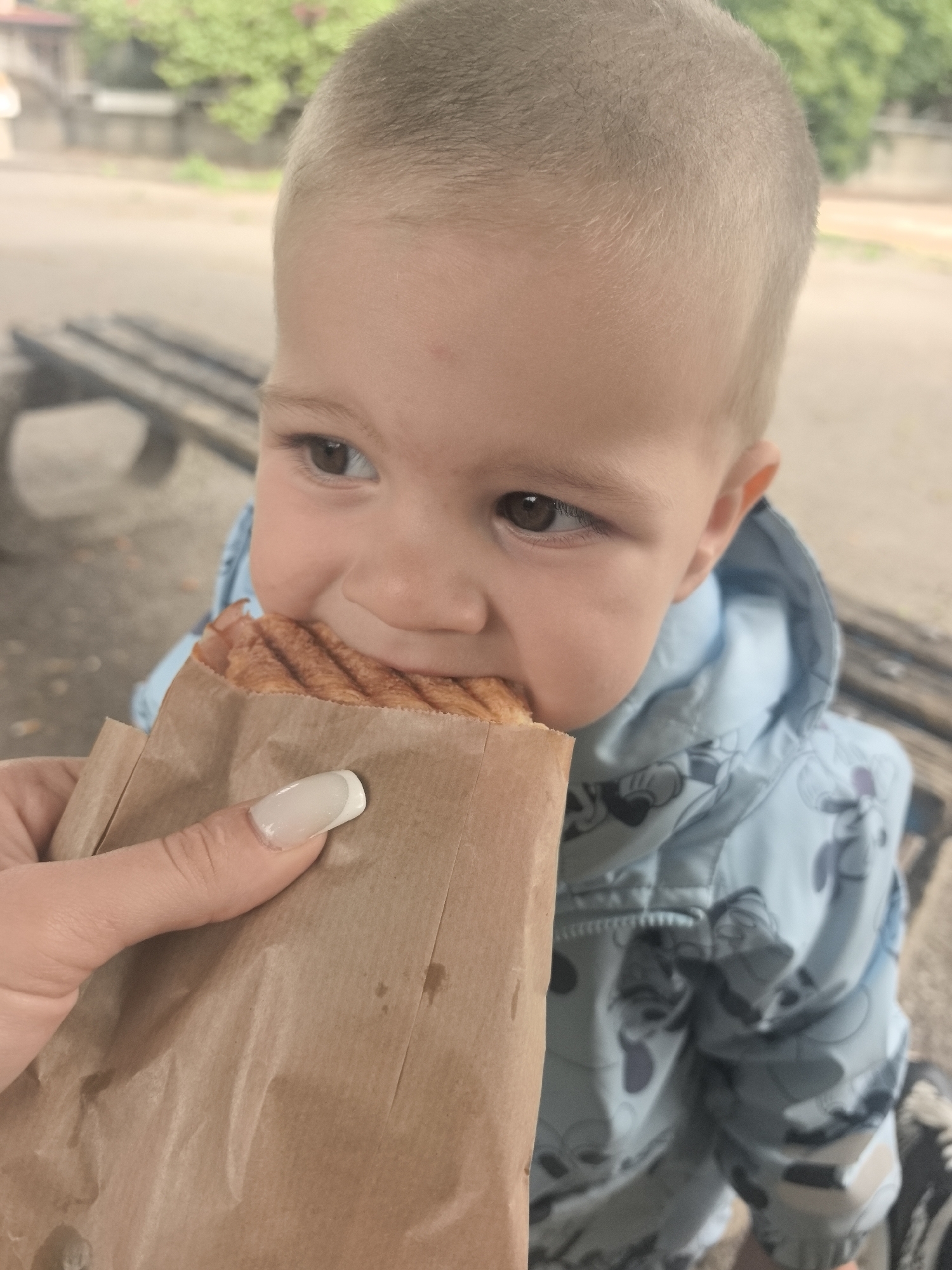 A young child with blonde hair is biting into a sandwich wrapped in brown paper. The child is wearing a light blue jacket with a cartoon pattern. A person's hand with a manicured nail is holding the sandwich for the child. In the background, there are wooden benches and greenery, suggesting an outdoor setting, possibly a park or public area in the city of Stara Zagora, Bulgaria. The lighting indicates it is daytime.