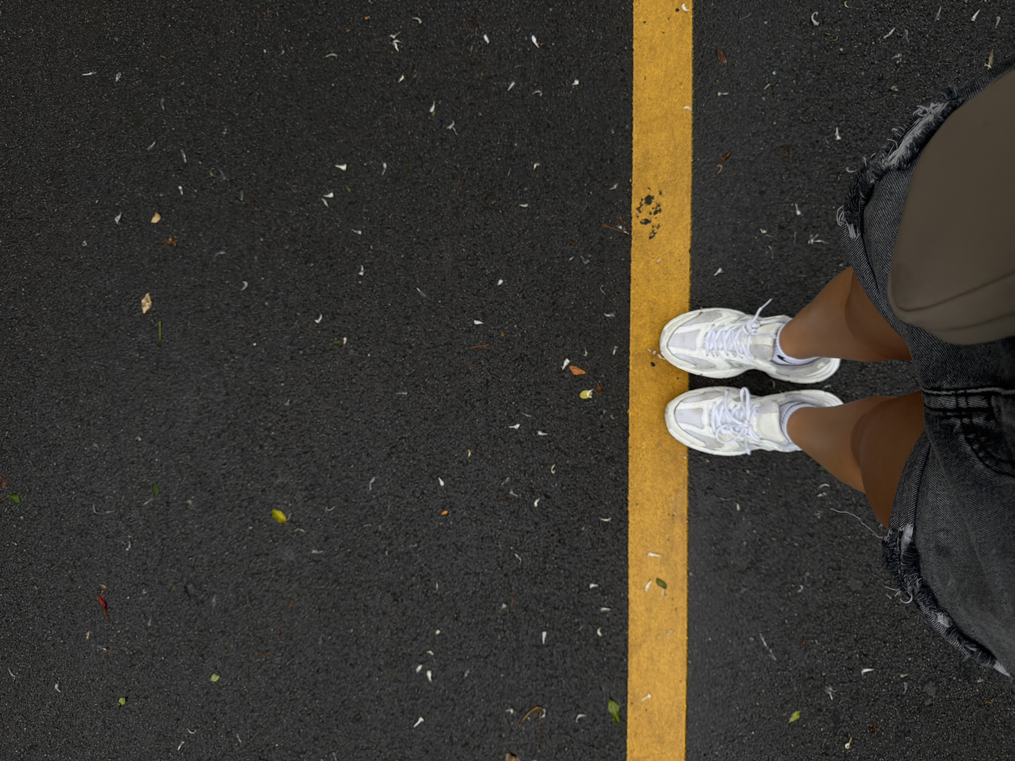 An overhead shot shows a person standing on a wet asphalt surface marked with a bright yellow line. The person is wearing white athletic shoes and dark denim shorts. There are scattered white petals and a few leaves on the ground. The scene appears to be an outdoor setting, possibly a street or walkway in Bangkok, Thailand, given the presence of scattered petals that are common from flowering trees in the region. The lighting suggests an overcast or rainy day.