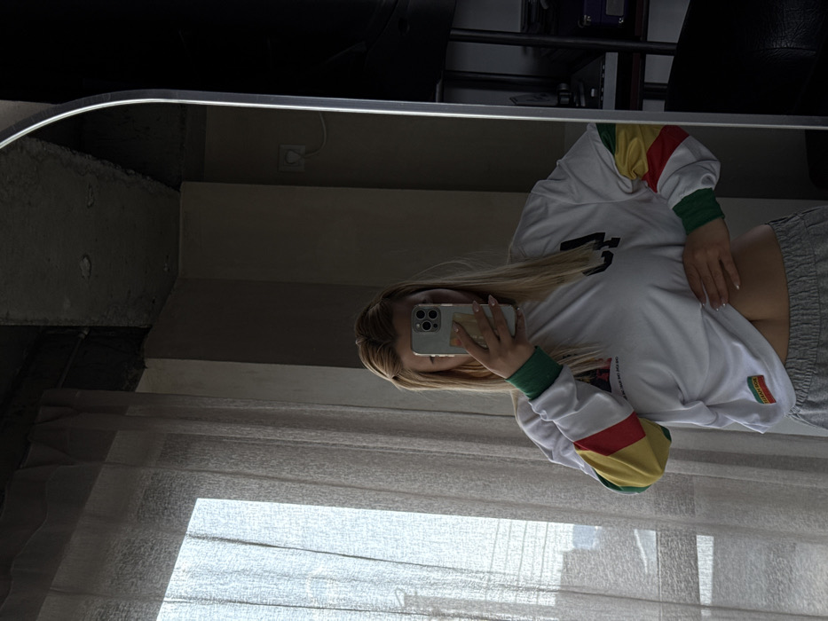 An individual with long blonde hair is depicted indoors taking a mirror selfie. The person's face is obscured by a light-colored smartphone held in their right hand. They wear a white long-sleeved shirt with multi-colored (green, yellow, red) stripes on the sleeves and grey sweatpants. The shirt is slightly lifted, exposing the abdomen. The indoor environment features a large window on the left, covered by sheer, light-colored curtains, showing a blurry urban background. The ceiling above the window and extending to the right is exposed, unfinished concrete. A light-colored wall with a white electrical socket is visible to the right. A tall, slender mirror with an illuminated edge reflects a dark furniture unit, possibly a shelving system or television stand, on the far right. The scene is illuminated by natural light entering from the window. The specific location within the city of Yerevan, Armenia, is not identifiable from the visual evidence.