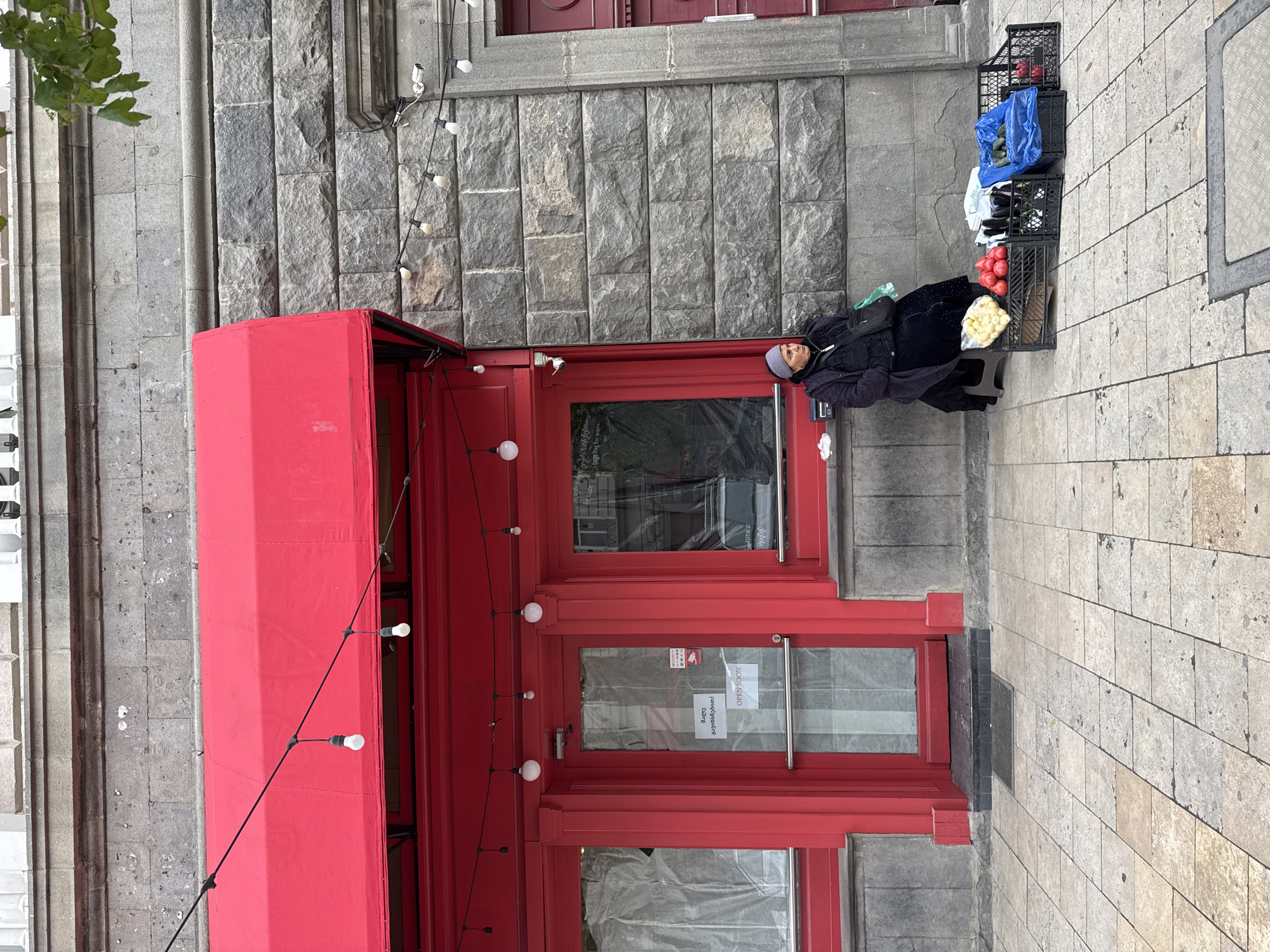 An outdoor scene in Tbilisi, Georgia, captures a street vendor selling fresh produce in front of a stone building with a vibrant red storefront. An older woman, dressed in a dark winter coat and a light grey beanie, sits on a low stool on the tiled pavement. She is positioned to the right of a display of goods.
Arranged on the pavement beside her are several black plastic crates filled with various fruits and vegetables. These include bright red tomatoes, dark purple eggplants, green cucumbers in a blue plastic bag, and a cluster of light yellow produce, possibly potatoes or lemons. A dark shoulder bag rests next to the vendor.
The building behind her features a facade of rough-hewn grey stone blocks. A prominent red awning extends over the storefront, which consists of red-framed doors and windows. A string of incandescent light bulbs is strung horizontally across the red awning and storefront. One of the red doors has a white sign with "OPEN SOON" in English and Georgian script ("მალე გაიხსნება!"). Green foliage from an unseen tree or bush is visible in the upper left corner. The scene is lit by natural daylight, suggesting it's daytime, likely with overcast skies given the soft light and lack of strong shadows. The overall impression is a bustling urban setting with a local market feel.