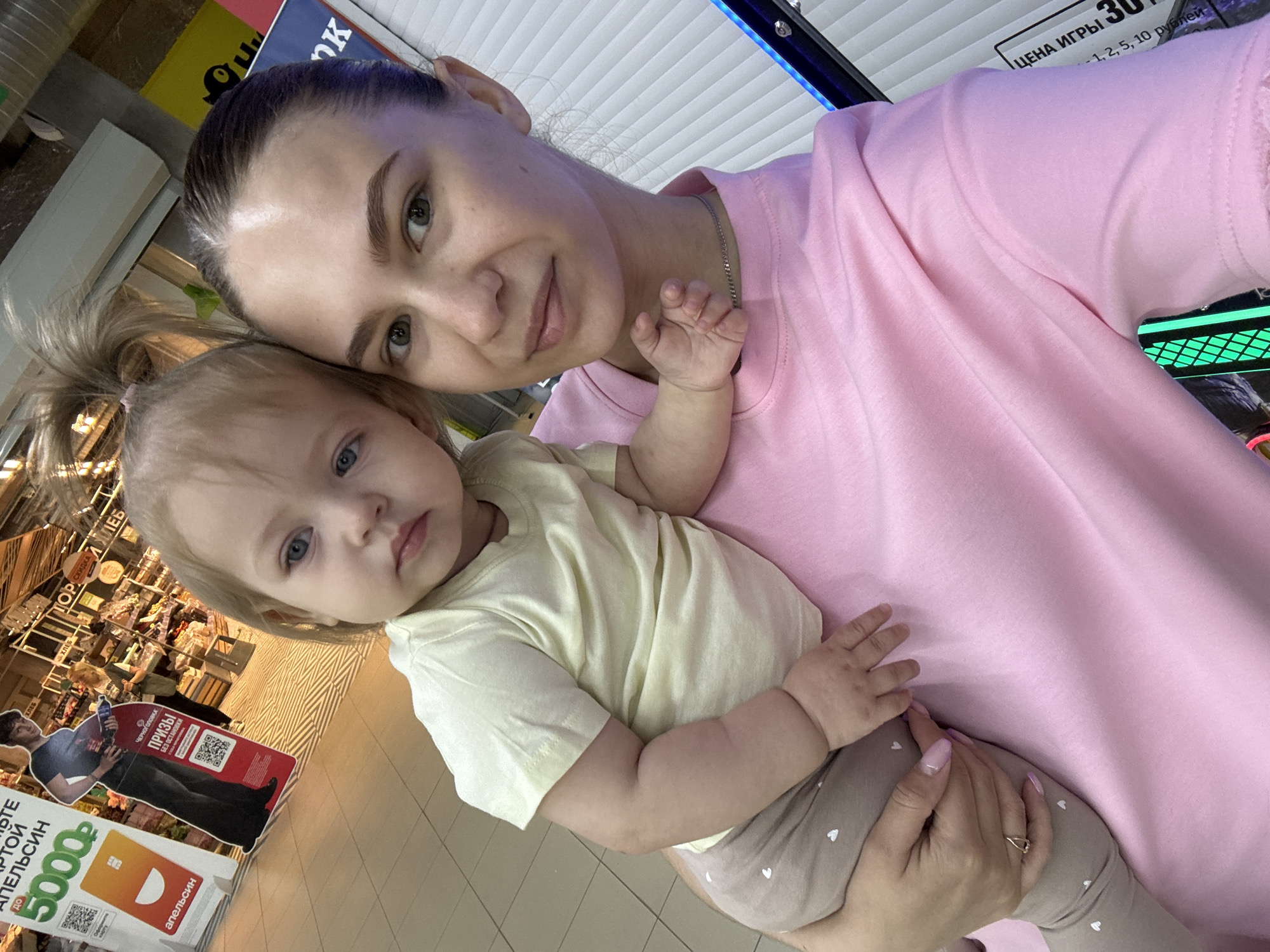 A selfie shows a woman and a baby. The woman is light-skinned with dark hair pulled back, wearing a pink t-shirt and a silver necklace. She is looking directly at the camera with a slight smile. The baby, also light-skinned with blonde hair, is wearing a light yellow t-shirt and light brown pants with white hearts printed on them. The baby is resting in the woman's arms, partially facing the camera. In the background, there are aisles of a store, and a promotional display stand on the floor for "Черноголовка" prizes, featuring an image of a man and a "5000" coupon. The sign also lists the city of Chaikovsky. Further in the background, there is a price list for a game; "ЦЕНА ИГРЫ 30 -1, 2, 5, 10 рублей" (Game Price 30 - 1, 2, 5, 10 rubles). The picture appears to have been taken in a store in Chaikovsky, Russia.