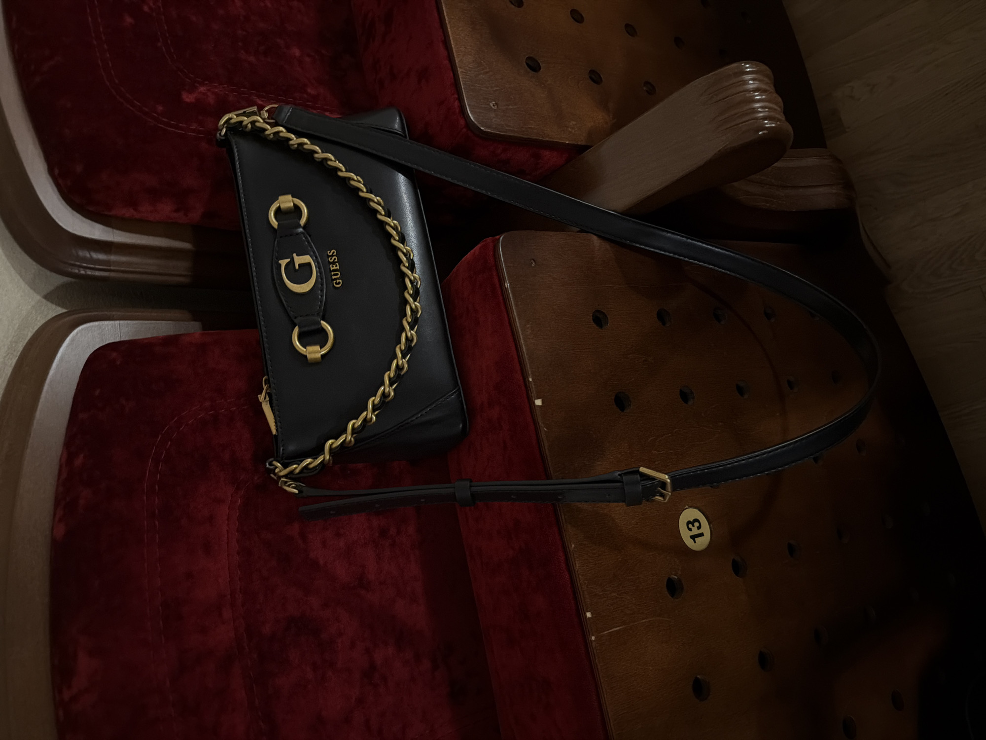 The image shows a black Guess purse with a gold chain strap and "G" logo lying on an old theater seat with red velvet upholstery. The seat is wooden, with rows of small holes and a "13" seat number plate. The setting suggests a vintage theater or cinema interior. It is impossible to verify the location in Izhevsk, Russia from the image itself.