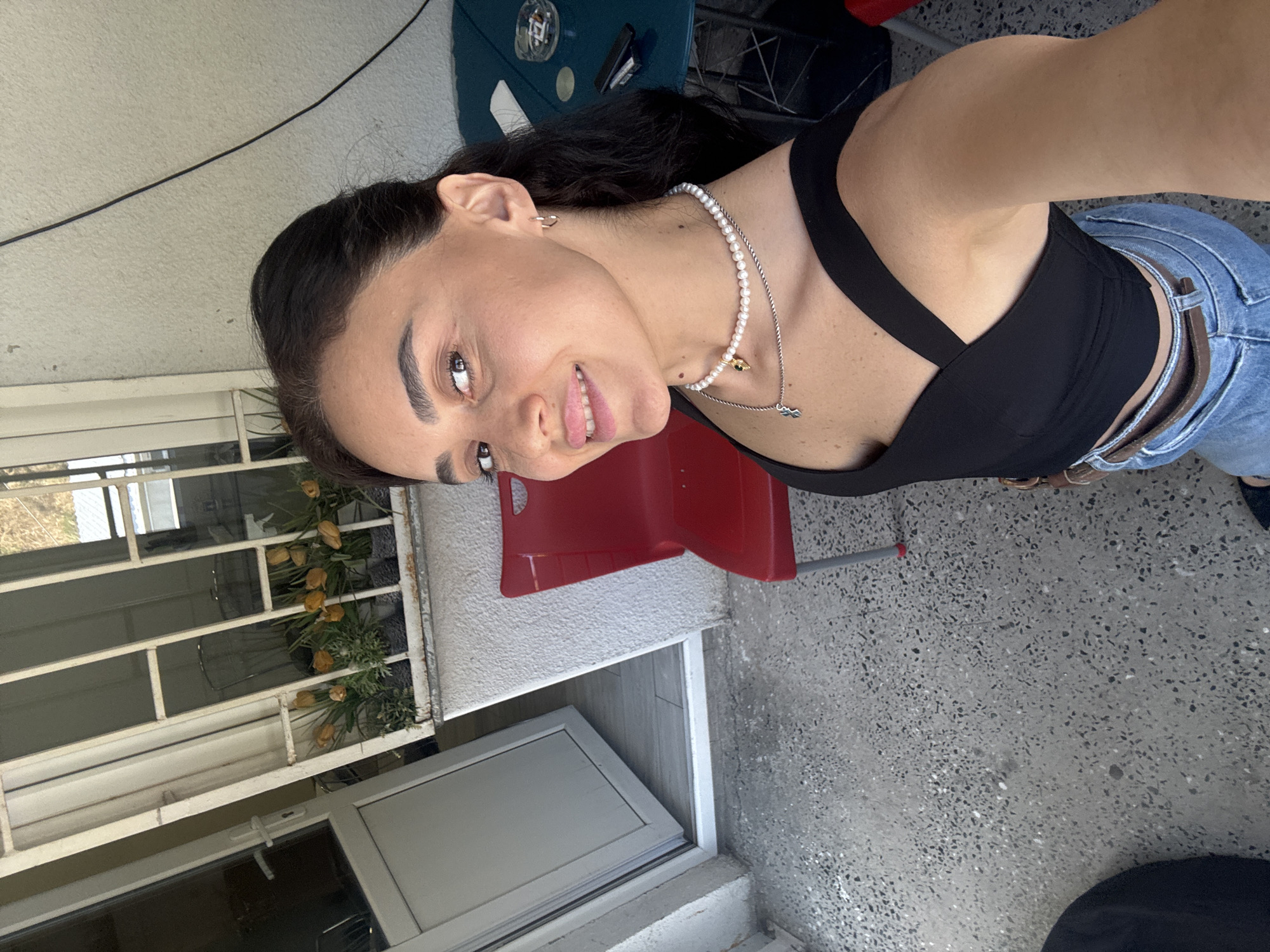 A young woman is taking a selfie on a balcony. She has dark hair and is wearing a black top and blue jeans. She is also wearing a pearl necklace and a pendant. In the background, there is a red chair, a table with an ashtray and cigarettes, and a door with a window. There are also some potted plants with yellow flowers on a ledge. The balcony floor is concrete. The image appears to have been taken in Tbilisi, Georgia, based on the architectural style and the general feel of the location.