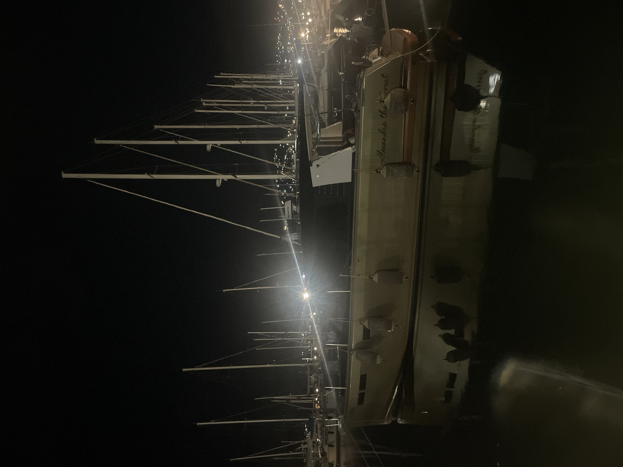 The image depicts a nighttime view of a marina in Bar, Montenegro, featuring several docked sailboats. In the foreground, a large sailboat named "Alexandra the Great" is prominently displayed. The boat is light-colored and reflects in the dark water. Three cylindrical fenders hang from the side of the boat. In the background, numerous other sailboats with tall masts are lined up, some with lights on. The distant shoreline is illuminated by city lights, suggesting the marina is located near an urban area. The sky is dark and clear.