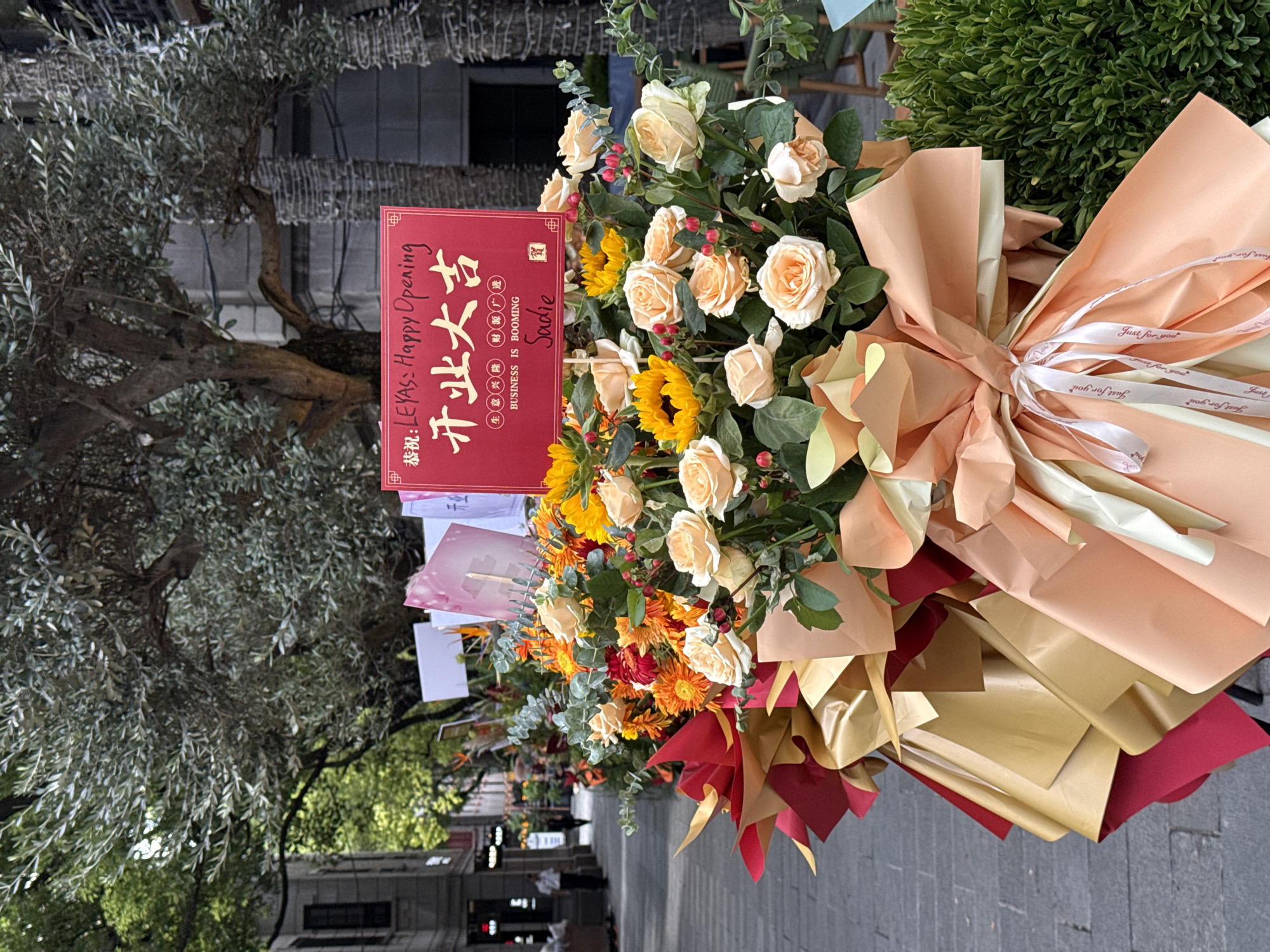 An outdoor, eye-level, medium shot captures a vibrant floral arrangement in front of a street. The arrangement features a variety of flowers in warm tones, including roses, sunflowers, and gerbera daisies, adorned with gold and red paper wrapping and bows. A red sign is prominently placed on the arrangement, with Chinese characters reading "开业大吉" (Happy Opening) and English text below that states "BUSINESS IS BOOMING Sadie". The background shows a tree with silvery-green leaves, a building with dark windows, and a cobblestone street. A few people are visible in the distance walking on the street. The overall scene suggests a celebratory occasion, likely an opening of a business, in Shanghai, China.