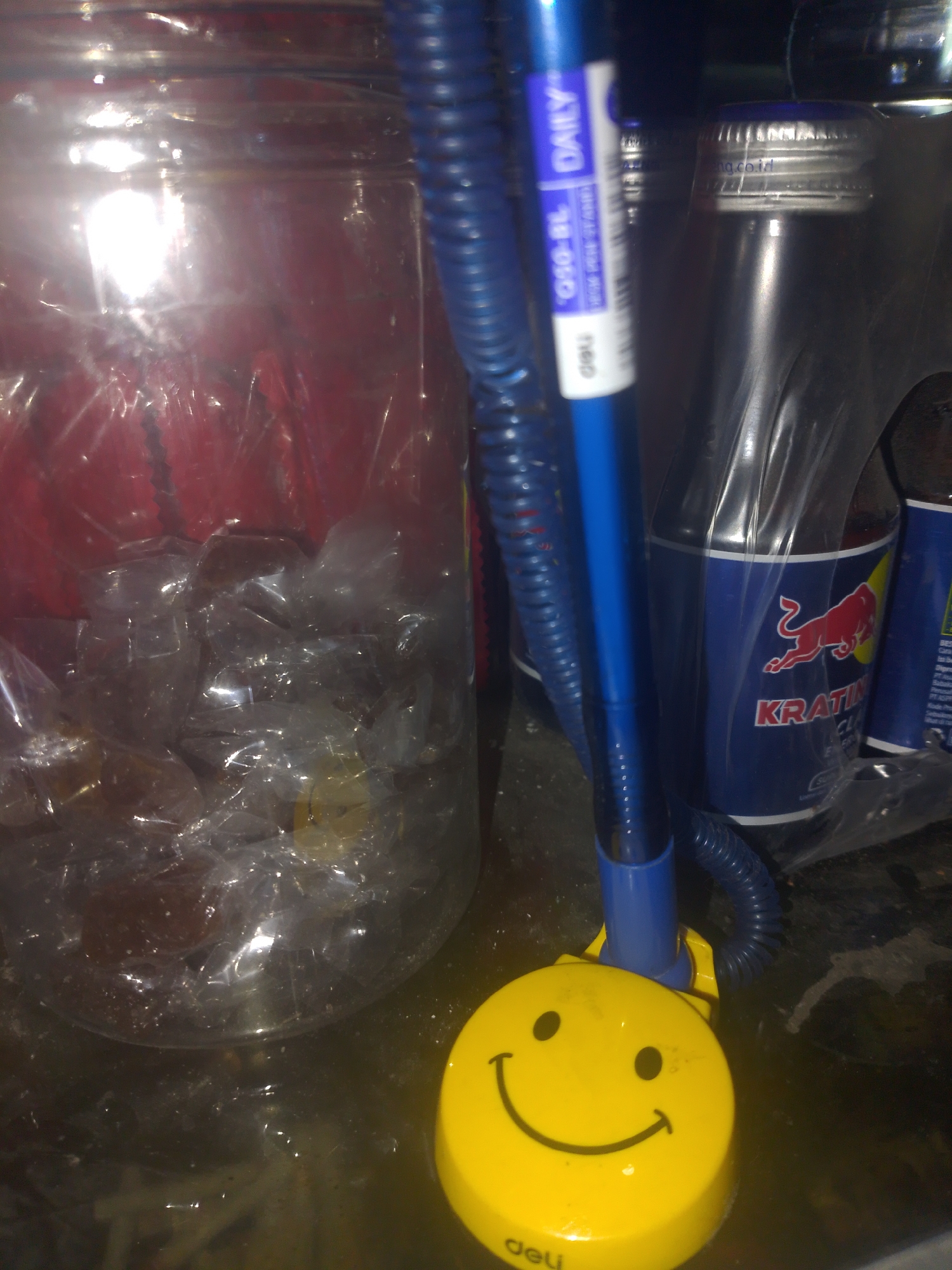 A close-up, slightly blurry image captures a collection of items on a dark, reflective surface. Dominating the foreground is a bright yellow, circular object with a smiling face emoticon, a common desk accessory. Attached to this is a blue plastic tube, which snakes out of frame to the right. Behind this, a clear plastic jar is filled with numerous individually wrapped, amber-colored candies. To the right of the jar, and partially obscured by the tube, are two dark blue bottles with silver caps. One bottle, clearly labeled with a red bull logo and the text "KRATINGDAENG", is the most discernible. The scene appears to be indoors, possibly in a store or home in Mukapayung, Indonesia, given the presence of local beverage brands. The lighting is dim, with a strong glare reflecting off the smooth surfaces.