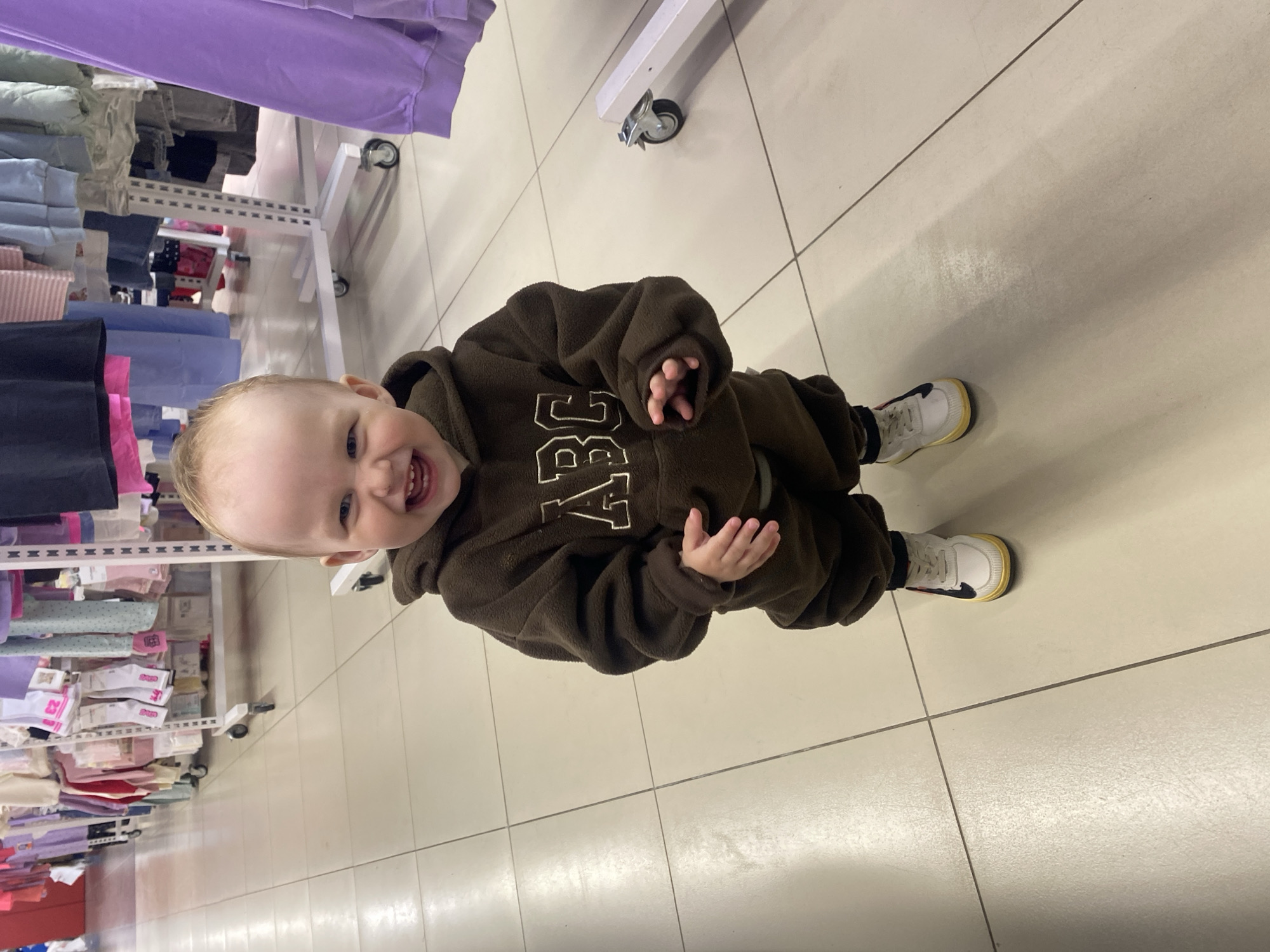A baby, around one year old, is standing on a light-colored, tiled floor inside a retail store. The baby is wearing a brown hooded jumpsuit with white "ABC" letters on the front and white sneakers with a yellow trim. The baby is smiling and looking up at the camera. Racks of folded clothes in various colors (purple, light green, blue, black, pink) are visible on the left side of the frame. The location is likely Chaikovsky, Russia, based on the prompt's instructions.