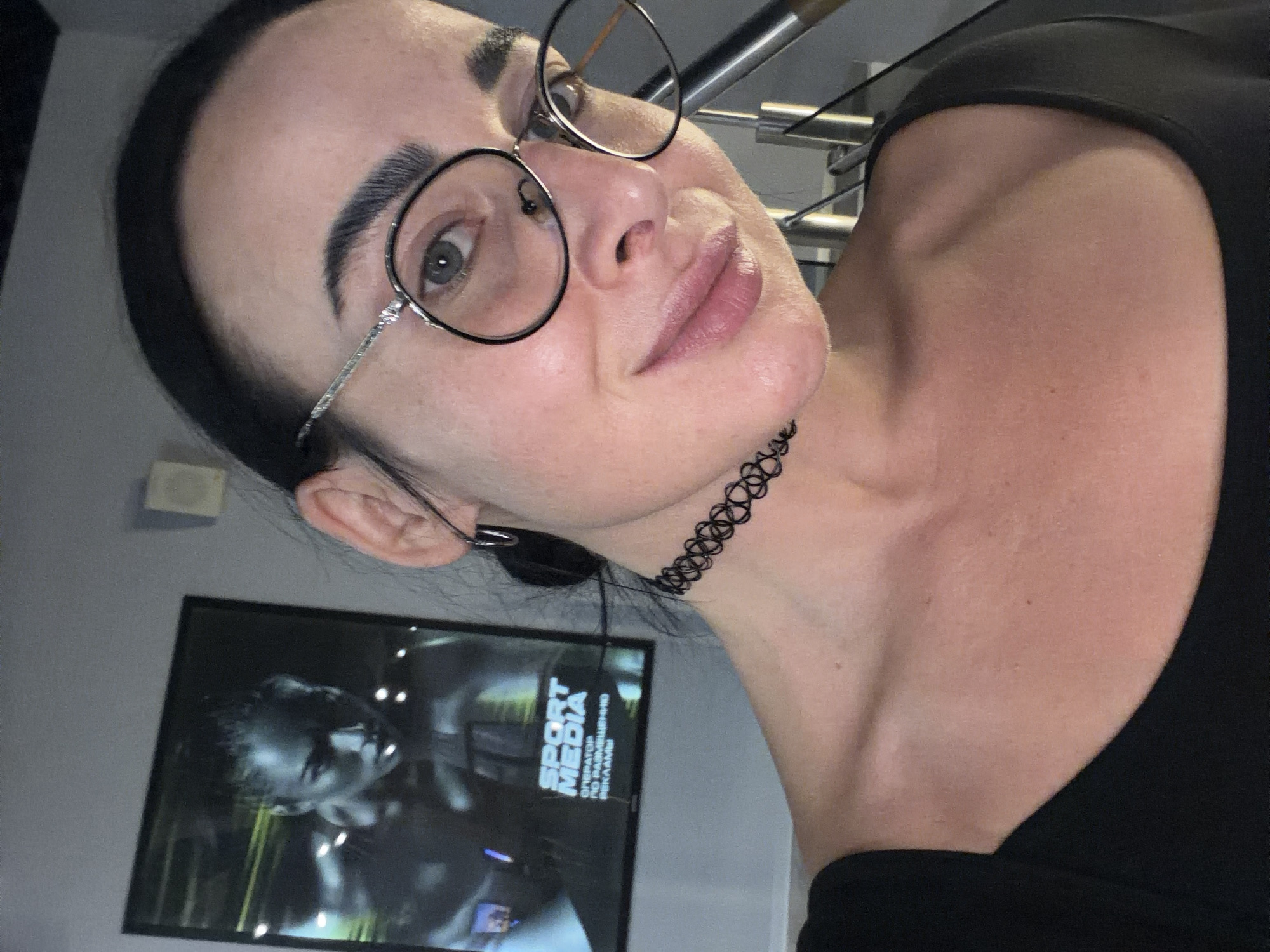 A woman with dark hair pulled back is wearing round glasses and a black choker necklace. She's wearing a black top. Behind her is a framed picture on a gray wall. The picture shows a person and the words "SPORT MEDIA" in Cyrillic. A set of stairs is visible in the background. It's not possible to confirm the location is Saint Petersburg, Russia from the given image.