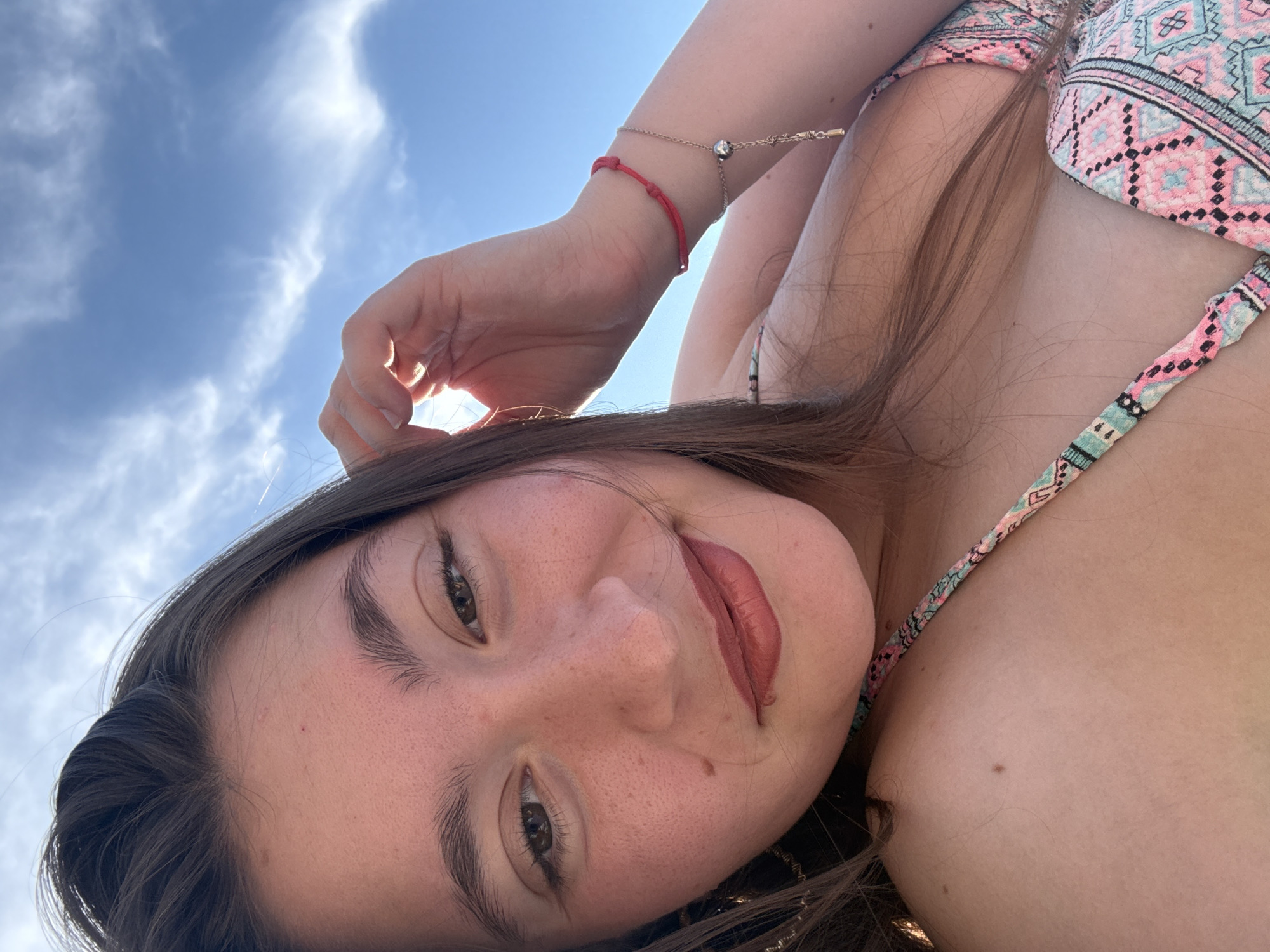 This is a close-up, low-angle selfie of a young woman against a bright blue sky with some wispy clouds. She is wearing a colorful bikini with a geometric pattern. Her hand is raised, touching her hair, and she has bracelets on her wrist, including a red string bracelet and a delicate gold chain bracelet with a small charm. She has long, brown hair, styled in loose waves. Her face is turned towards the camera, with a soft smile and makeup on her lips. The lighting suggests it is daytime, likely sunny given the bright sky. The image conveys a sense of relaxation and enjoyment, possibly taken outdoors on a warm day. The location context indicates this may have been taken in Sozopol, Bulgaria. There is no visible text in the image.