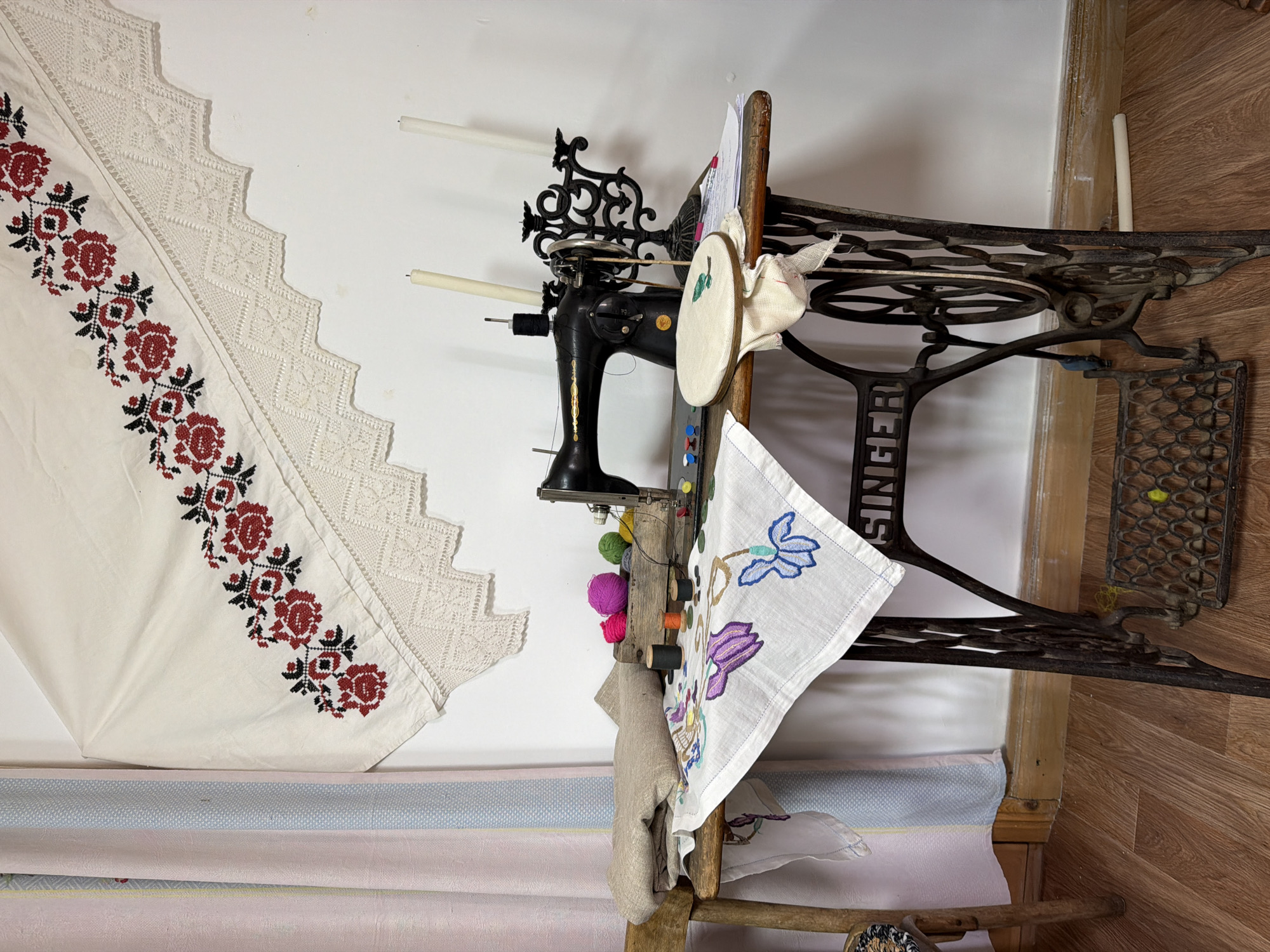 The image shows a close-up view of a sewing machine and surrounding crafting materials on a wooden table. The sewing machine is black and appears to be an antique Singer model. Various spools of thread, skeins of colorful yarn, and buttons are scattered on the table. A piece of white fabric with embroidered blue flowers is draped over the edge of the table, and another piece of fabric, folded, is on a wooden chair to the left. Above the sewing machine, a white textile with red floral embroidery and lace trim is hung on the wall. Two white candles in a decorative black candelabra stand behind the sewing machine. The scene is set against a plain white wall and a wooden floor. The overall impression is of a crafting or sewing area, possibly a workspace or a display in a home or small shop. The specific location in Ust'-Kabyrza, Russia is not determinable from the image itself.