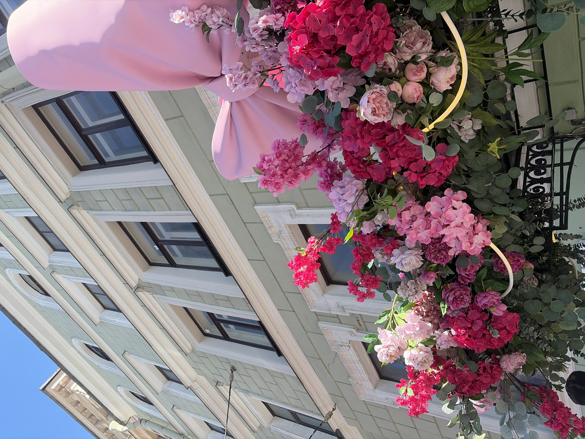 An exterior, low-angle shot captures a building facade adorned with a large, pink fabric bow and an abundant floral arrangement. The building, painted a pale green, features multiple windows with dark frames and white trim. The flowers are a vibrant mix of pink and fuchsia hydrangeas and peonies, interspersed with green foliage. The arrangement is suspended above a dark, decorative metal structure. The sky above is clear and blue.