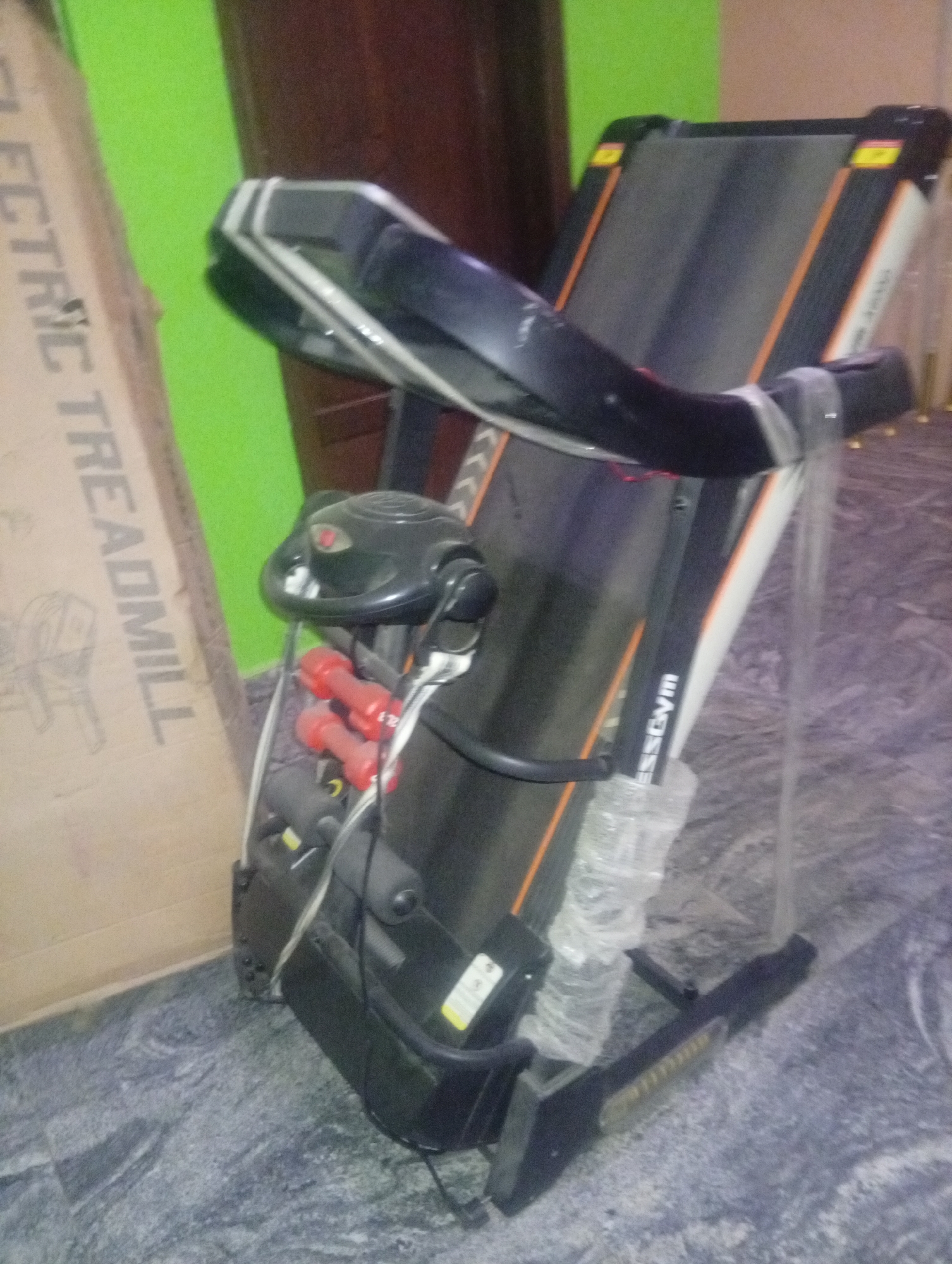 The image depicts a folded treadmill in an indoor setting, likely a residence. The treadmill is black with orange and white accents, bearing the word "FitnessGym" on the side. Two small red dumbbells are attached to the console area. Protective plastic wrapping partially covers the machine's frame. A cardboard box labeled "ELECTRIC TREADMILL" is visible to the left. The background includes a green wall and a brown wooden object. The floor has a gray, patterned surface. The location is Aviation Quaters, Nigeria. No people are visible.