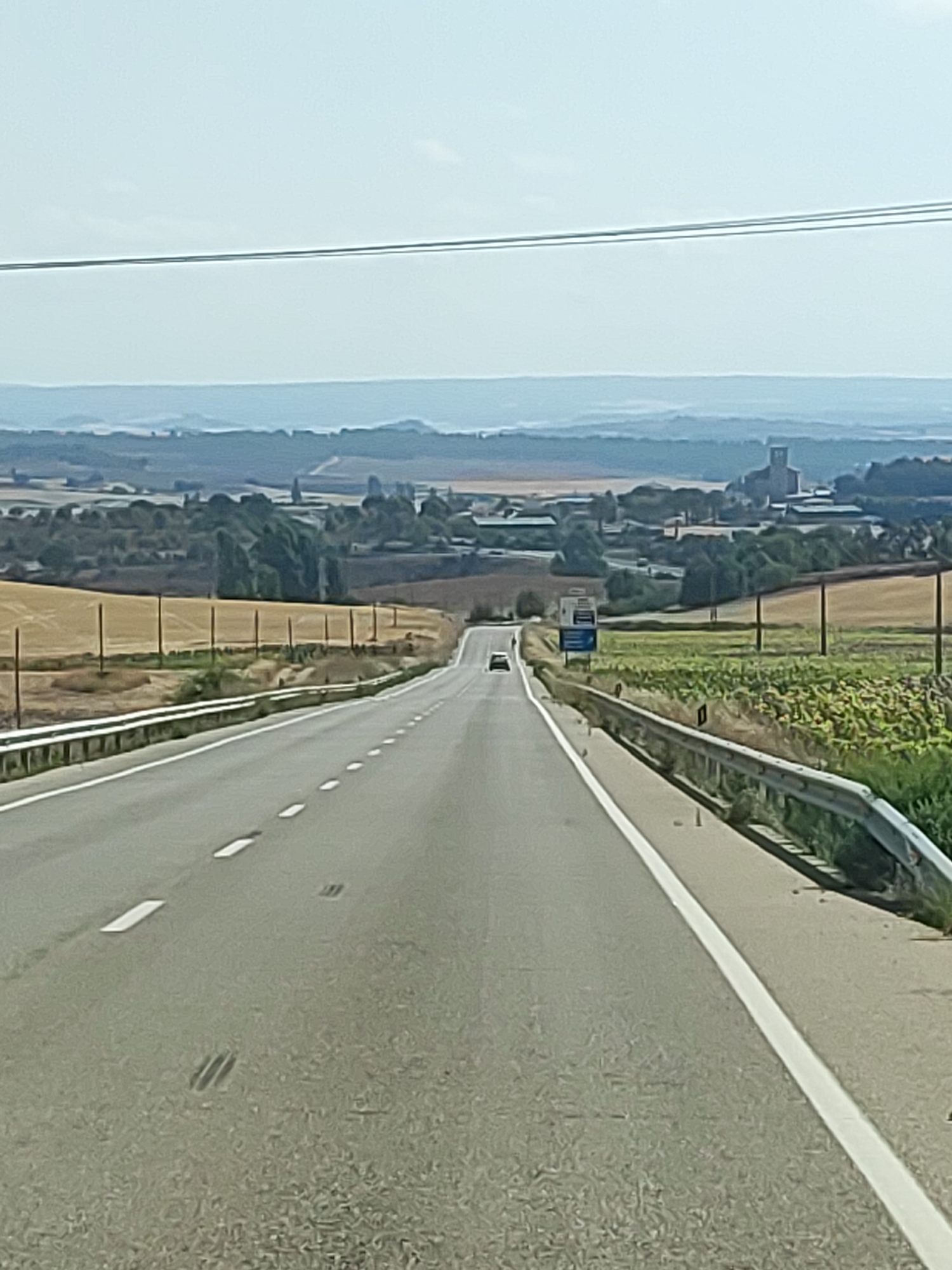 A photograph captures a view from a vehicle traveling on a straight, asphalt highway. The highway has a white solid line on the right edge and dashed white lines in the center. A metal guardrail runs along the right side of the road, bordering fields of green crops. In the distance, the landscape is dotted with trees and some structures, including a prominent building that appears to be a church or tower. A hazy sky hangs over the scene. The image was taken in La Almarcha, Spain.