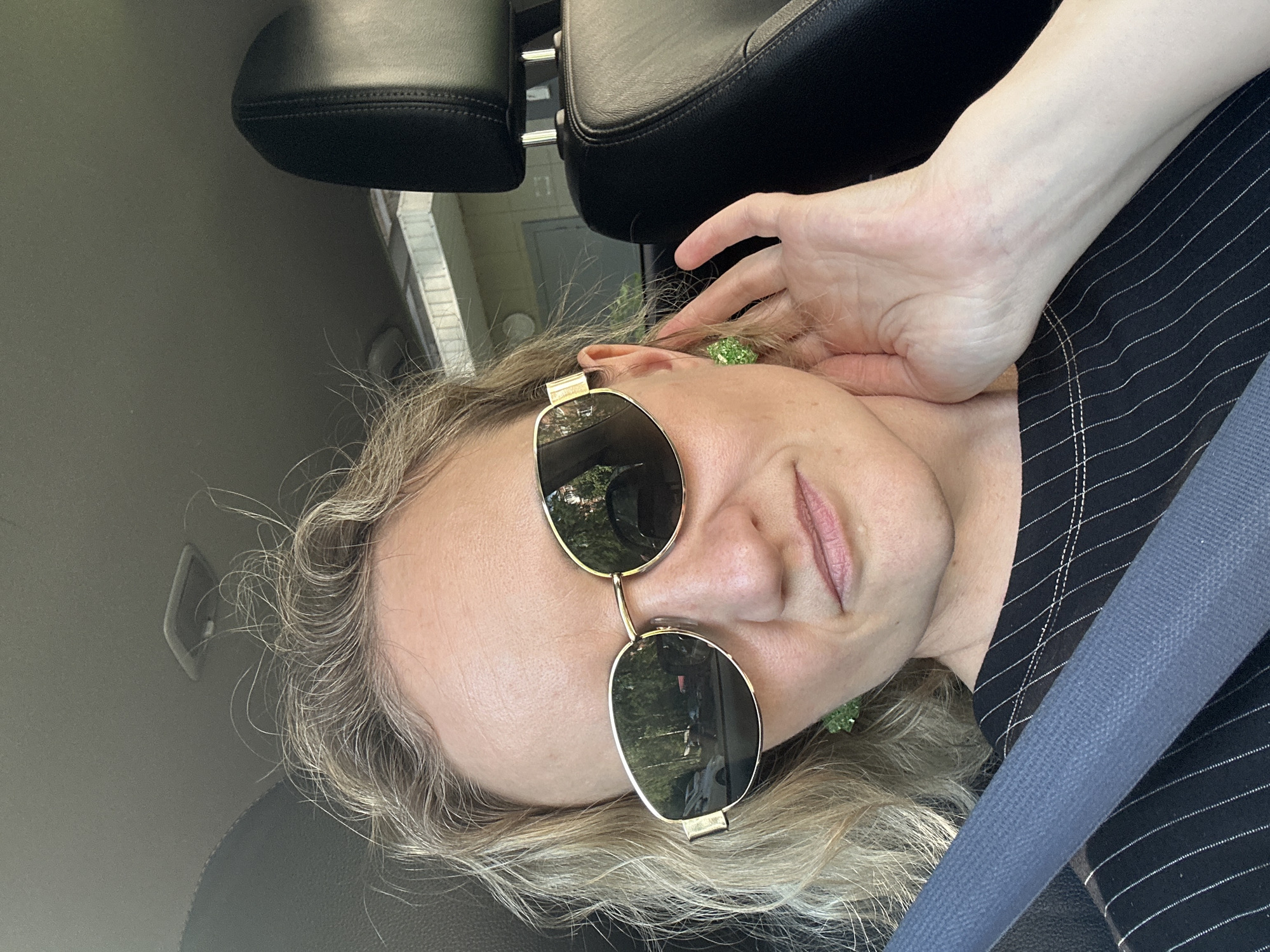 A woman is taking a selfie inside a car. She is wearing sunglasses and a pinstripe shirt and a gray seatbelt. Her hand is near her ear, revealing a green earring. The car's headrest is visible in the background. The location is indiscernible.