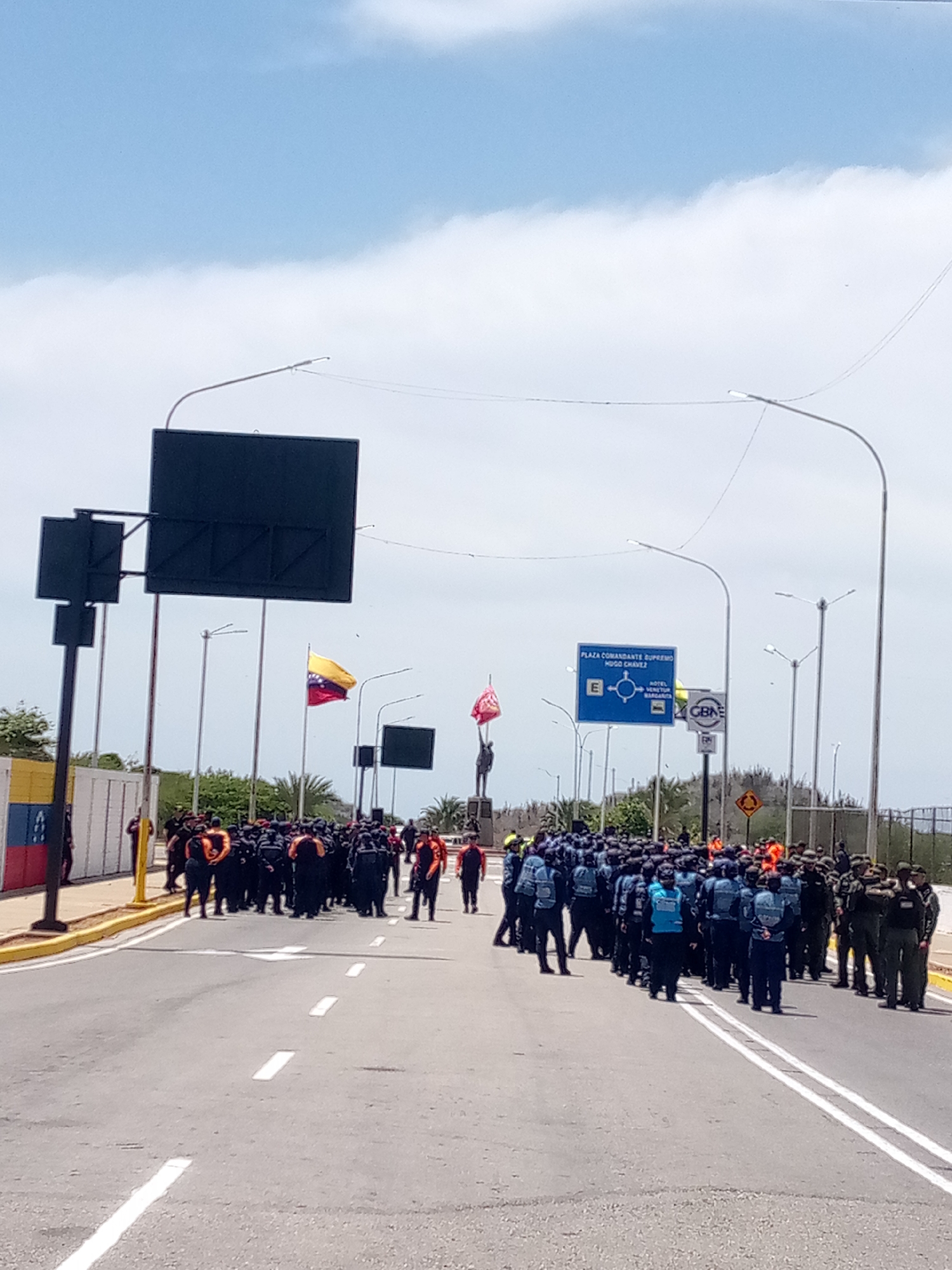 The image depicts a street scene in Porlamar, Venezuela. A large group of people, some in dark uniforms with orange accents and others in blue uniforms, are standing on a road. Some individuals appear to be in military-style attire. A statue is visible in the background, along with Venezuelan flags. Roadside signs, including one that reads "Plaza Comandante Supremo Hugo Chavez," are also visible. The sky is partly cloudy.