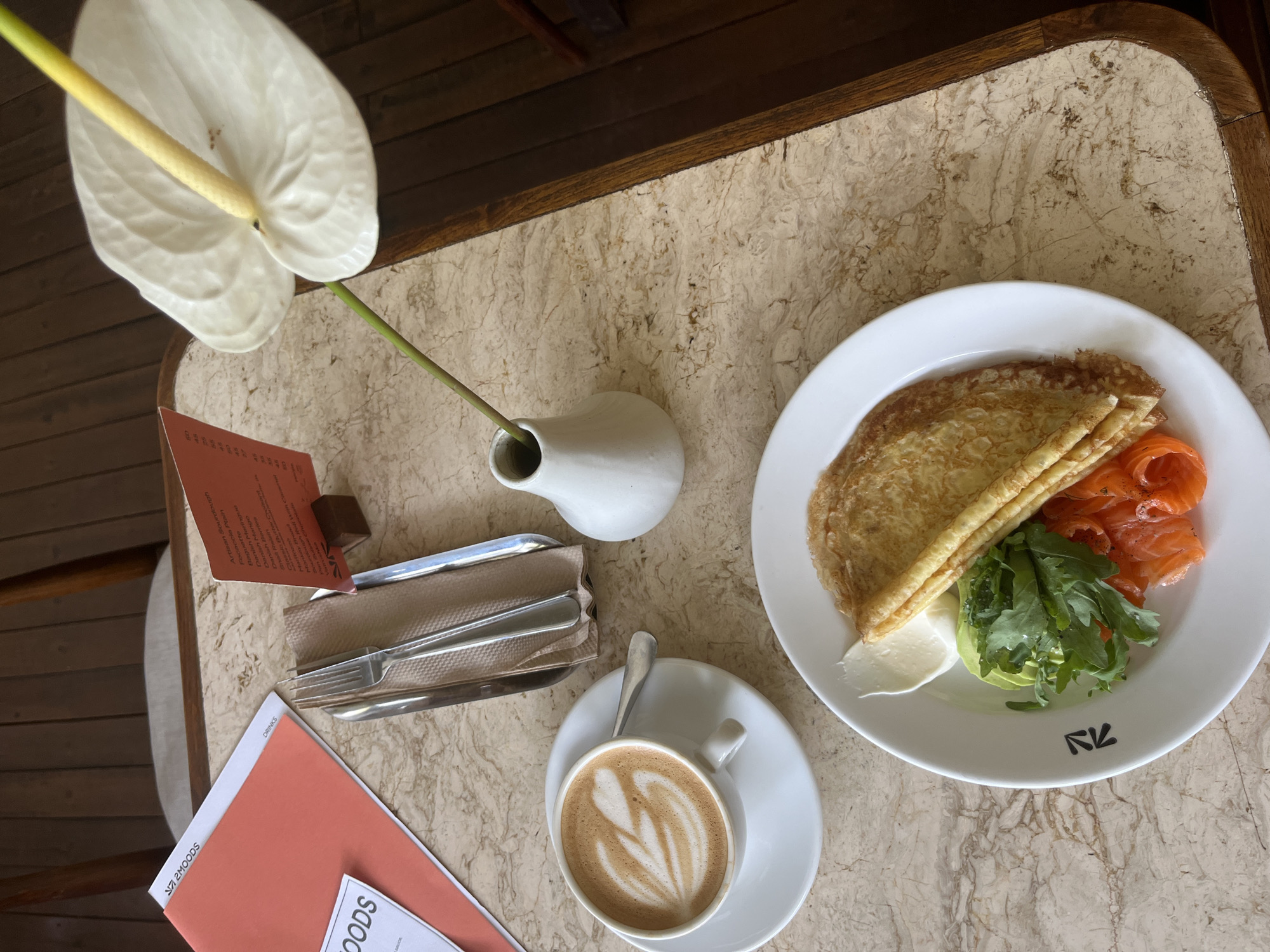 The image shows a tabletop scene, likely captured at a cafe in North Kuta, Indonesia, given the menu indicating '2MOODS' and the type of cuisine. A white Anthurium flower stands in a small white vase, positioned near a menu, silverware, and a coral-colored card. A white plate contains a crepe, accompanied by sliced salmon, avocado, and green leafy vegetables. A cappuccino is set beside the plate on a matching white saucer. The table has a beige marble-patterned top with a wooden edge.
