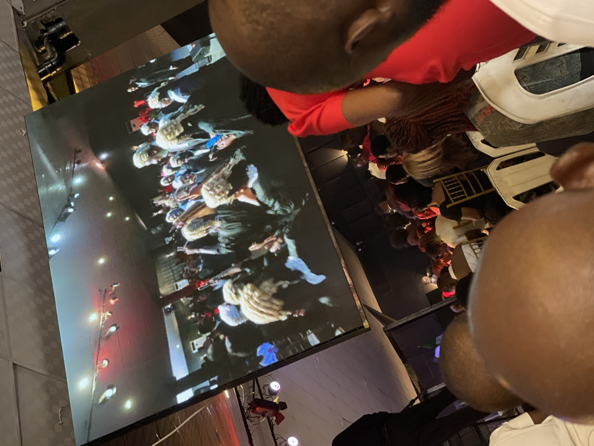 The scene appears to be indoors in a venue or event space in Abuja, Nigeria. A large LED screen displays what looks like a parade or public event with many people in costumes or uniforms. A crowd of people is gathered to watch, although their faces are mostly obscured or out of focus. A person wearing a red shirt is prominent in the foreground, seemingly close to the screen. Several white plastic chairs are visible. The overall lighting is dim except for the bright display on the screen.