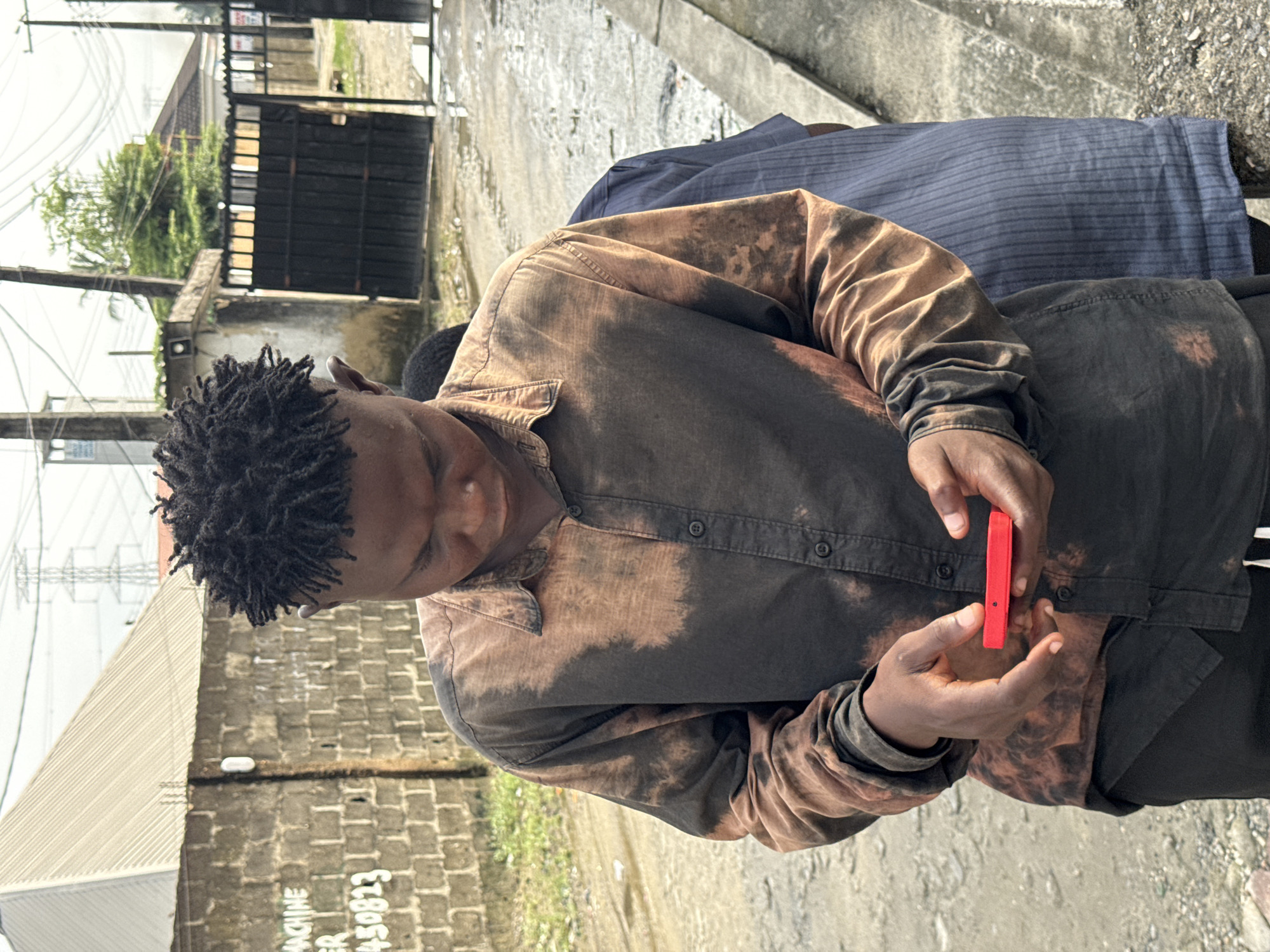 An outdoor image, rotated 90 degrees clockwise, captures a street scene in Ado, Nigeria, under bright daylight.
The primary subject is a person, likely male, with dark skin and dreadlocks, wearing a dark brown long-sleeved shirt with distinctive lighter brown, tie-dye or bleach-stained patterns. He is looking downwards, focused on a small, vibrant red, rectangular object held in his hands. This object, possibly a phone, vape pen, or small electronic device, has a tiny black dot or lens on one side. Behind him, another person is partially visible, wearing a dark blue striped shirt or garment.
The setting appears to be an urban or suburban environment. The ground is a mix of concrete and possibly paved areas. The background features various structures, including concrete or brick walls, dark metal gates and fences, and green trees and bushes. Utility poles and overhead wires are visible against the bright sky, suggesting a typical street or pathway. On a concrete block or wall in the lower left of the frame, partially obscured text reads "MACHINE" and "ER 450823". The overall lighting indicates a clear day with strong natural light. The main subject's expression is neutral, indicating concentration on the object he is holding.
