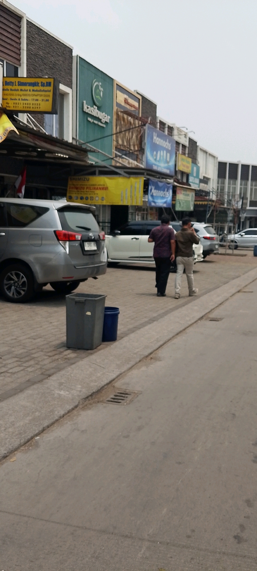 This is a medium shot of a street scene in Cileungsi, Indonesia, captured from a low angle. In the foreground, a grey garbage bin and a blue bucket are placed on a paved sidewalk next to a drainage grate. A silver SUV is parked on the left side of the image, partially obscuring the sidewalk. Further down the street, two men are walking away from the camera, one in a maroon shirt and dark pants, the other in a tan shirt and light-colored pants. They are walking on the sidewalk in front of a row of buildings that appear to be commercial establishments. The buildings have signs, including one that reads "kasibugar" in green text, and another with "Hannochs" in blue. A yellow banner with the word "SHIMIZU" is also visible. A white car is parked in front of one of the buildings. The sky is overcast and the overall lighting suggests it is daytime.