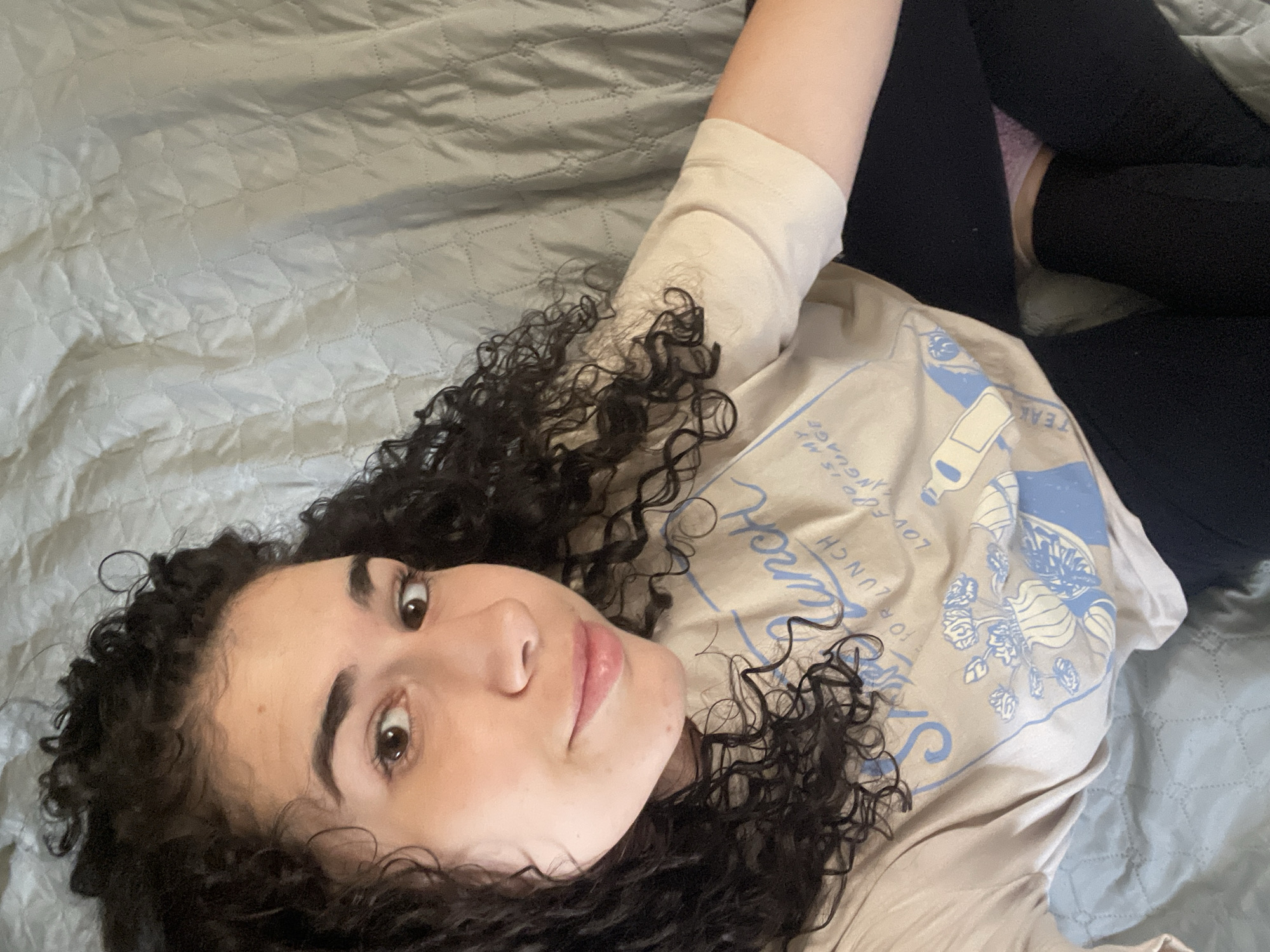 A young woman with curly dark hair is lying on a light blue quilted bedspread. She wears a tan t-shirt with blue print and black leggings. Her face is angled towards the camera, and she appears to be looking directly at the viewer. The photo offers no evidence of the location being Sofia, Bulgaria.