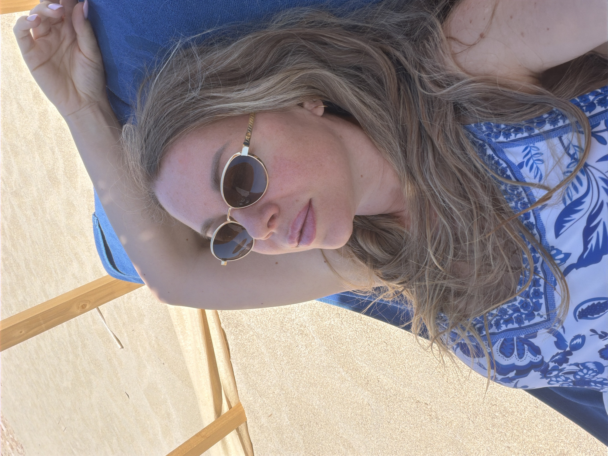 A medium shot captures a blonde woman lounging on a blue beach chair on a sandy beach, her face turned upward and slightly to the left, wearing sunglasses and a blue and white patterned swimsuit. The background is dominated by light brown sand and the wooden frame of the beach chair. The woman's arm is raised behind her head, and her hair is long and wavy, falling across her shoulder and chest. Her lips are slightly parted, and her eyes are hidden behind her sunglasses. The overall lighting suggests a sunny day.