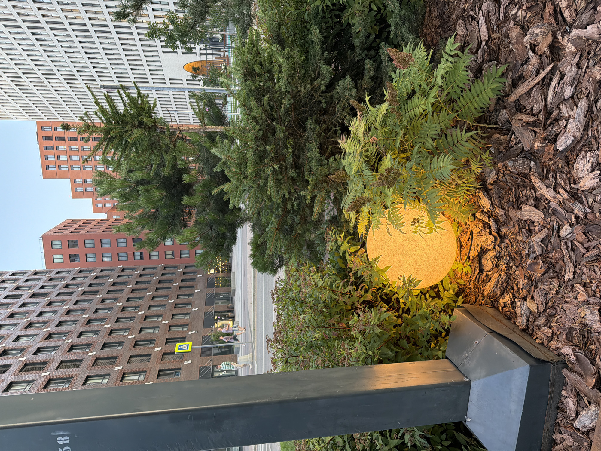 The image shows a globe-shaped light fixture, illuminated with a warm yellow glow, nestled amongst dense green foliage and bark mulch. The light is positioned near the base of a dark grey, angular pillar, possibly a street lamp or part of an outdoor installation. In the background, several modern, multi-story buildings with brick and white facades dominate the cityscape. The architecture suggests an urban environment, likely Moscow, Russia, characterized by contemporary residential or office complexes. A street with a pedestrian crossing is visible, with a few individuals walking on the sidewalk. The sky is a clear, pale blue, indicating daytime, possibly late afternoon or early evening given the artificial lighting.