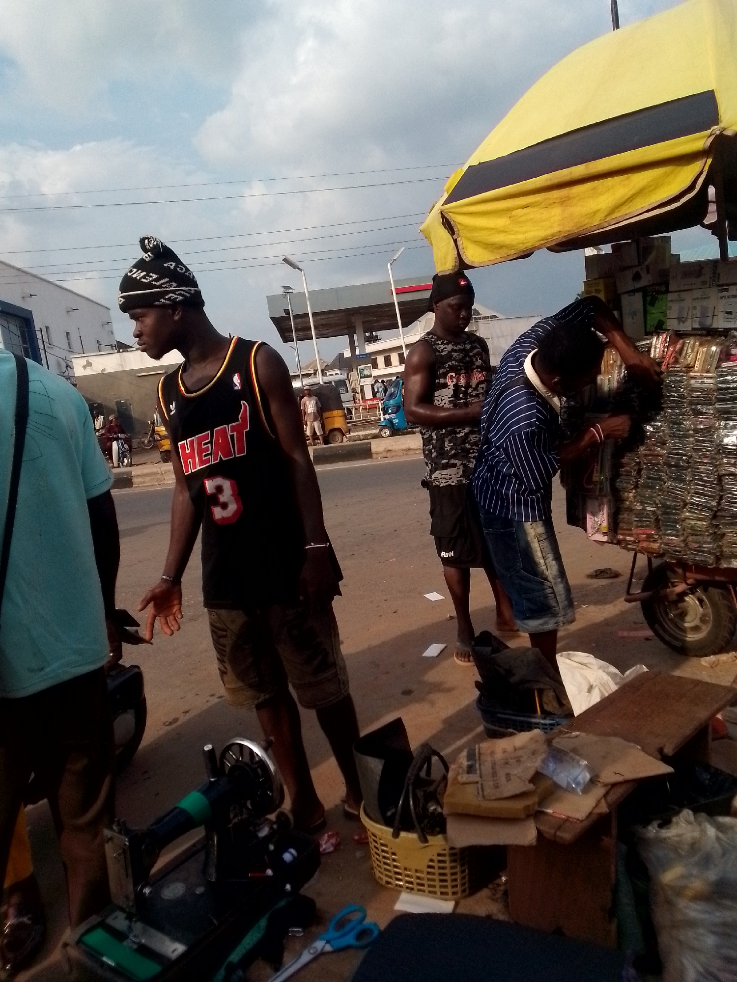 The image captures a street scene in Orlu, Nigeria. Three men are visible: one in a "HEAT 3" basketball jersey and beanie, another in a camouflage tank top, and a third in a blue striped shirt. The man in the striped shirt is interacting with a cart piled high with merchandise, likely phone cases or similar small items. The man in the HEAT jersey stands near a sewing machine, suggesting a tailoring business. The man in the tank top stands behind him. Debris, including boxes, baskets, and cloth, clutter the ground. In the background is a petrol station, and a three-wheeled vehicle "Keke" is visible. The sky is overcast.