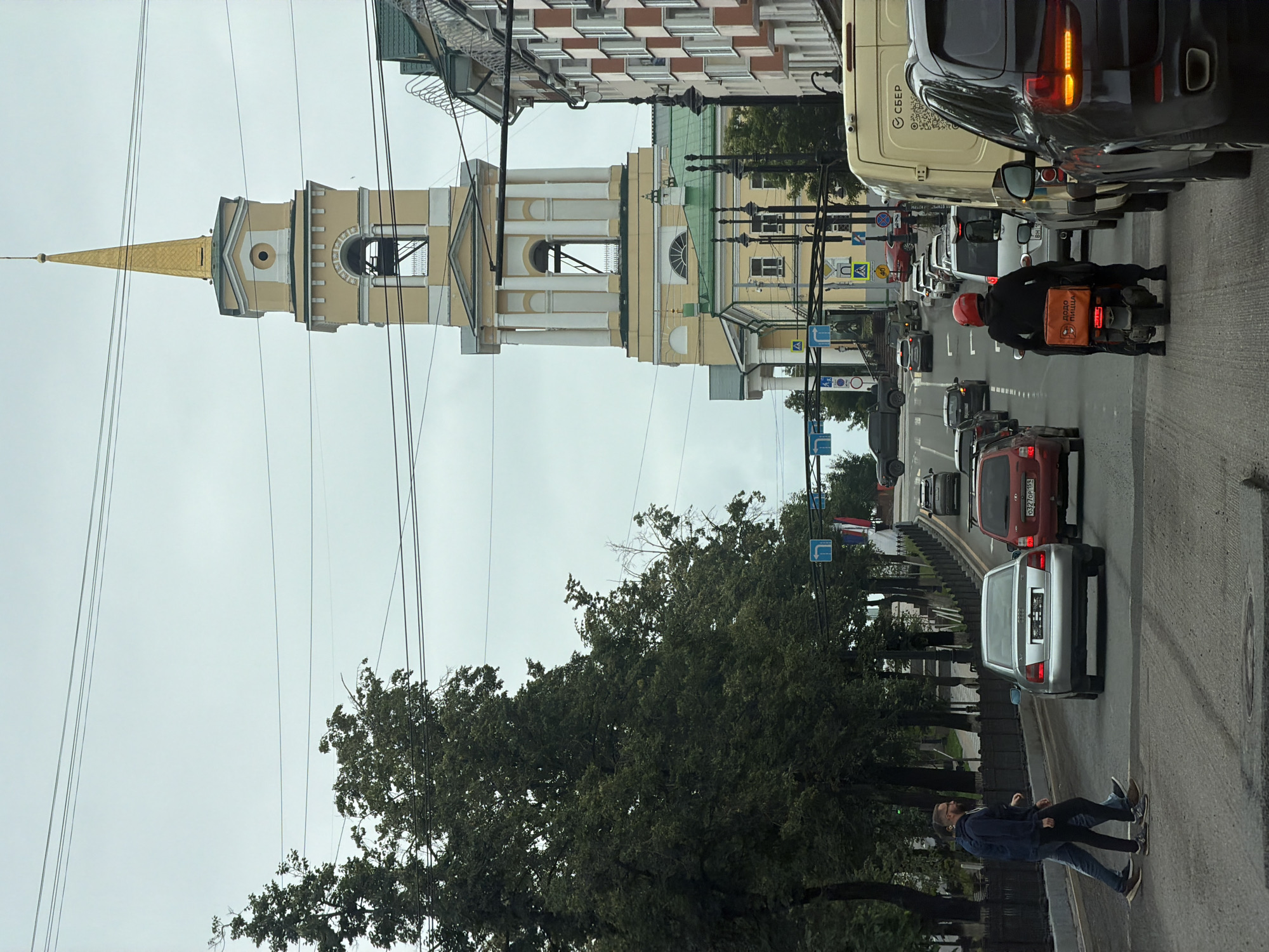 The image shows a street scene in the city of Perm, Russia. A tall, yellow and white church tower with a gold spire dominates the background. The church appears to be an Orthodox church. The street is busy with traffic, including cars, a scooter, and a delivery van. Two pedestrians are crossing the street in the foreground. The weather is overcast. The scene appears to be on a slightly sloped street, with trees lining the left side and buildings on the right. Traffic signs are visible, indicating traffic flow and pedestrian crossings. The delivery van has Cyrillic text on its side, which reads "СБЕР", indicating it's likely associated with Sberbank, a major Russian bank. The scooter is equipped with a thermal delivery bag that says "DODO PIZZA" in Cyrillic, indicating a food delivery service.