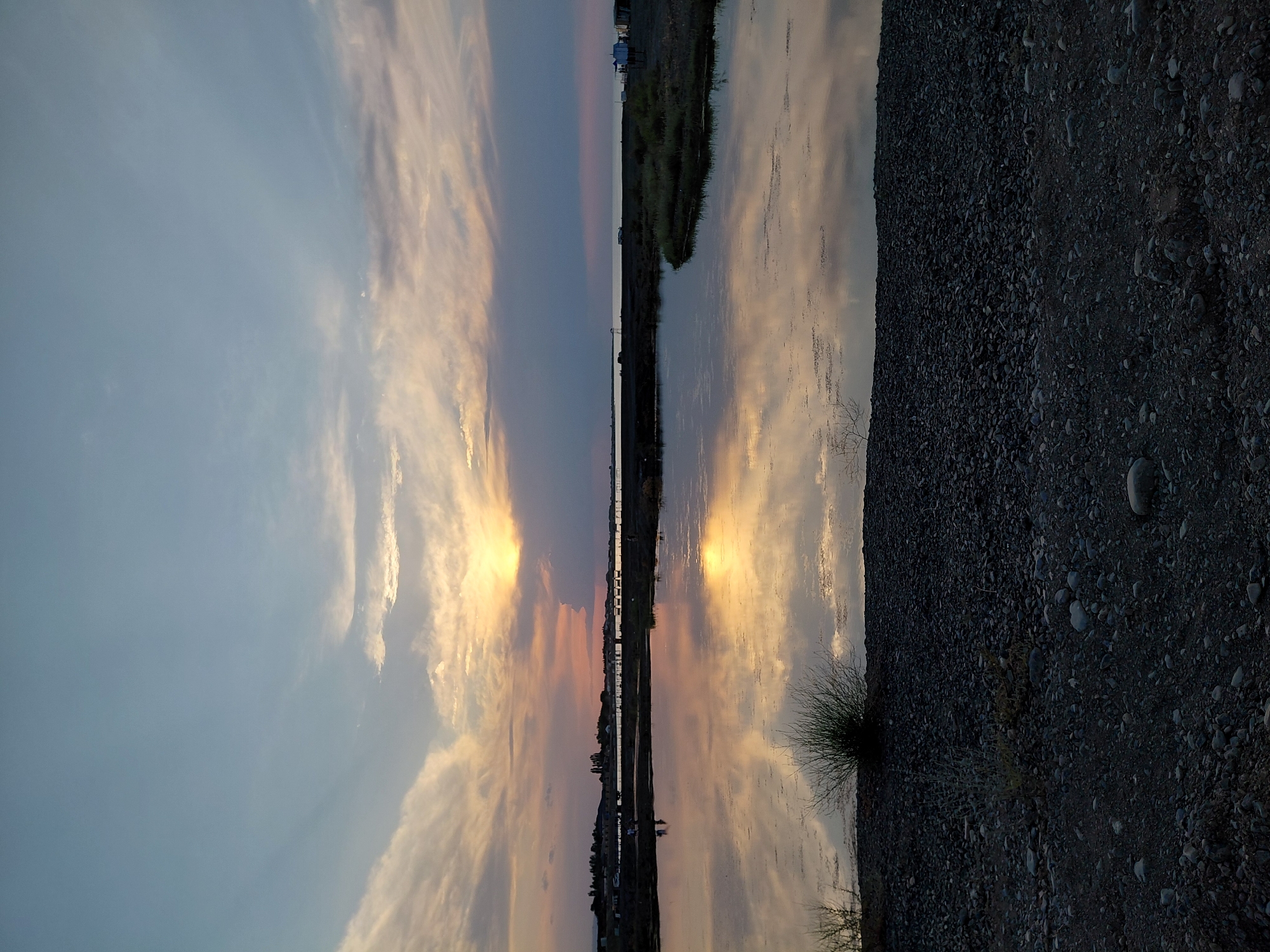 This is a landscape photograph taken at sunset in Akshi, Kazakhstan. The image is oriented vertically. The scene depicts a body of water, likely a river or canal, reflecting the sky and the surrounding landscape. On the left side of the image is the sky, filled with wispy clouds tinged with the warm colors of sunset – oranges, yellows, and pinks. These clouds are mirrored in the still water, creating a symmetrical effect.
The right side of the image is dominated by a gravelly shore, sparsely dotted with small rocks and some dry vegetation. In the distance, across the water, is a low-lying landscape with some scattered buildings and what appears to be a power line. The time of day is clearly sunset, indicated by the dramatic colors in the sky and their reflection. The weather appears calm, with still water suggesting little to no wind. The overall emotion evoked is one of tranquility and the beauty of a natural sunset. There is no visible text in the image.