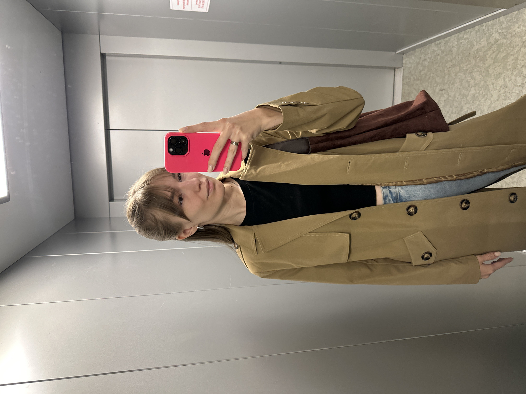 A woman is taking a selfie in what appears to be an elevator, based on the smooth, metallic walls. She is holding a bright pink smartphone up in front of her face. She is wearing a black top, blue jeans, and a light brown trench coat. A brown bag is slung over her shoulder. The location is likely within a building in the city of Chaikovsky, Russia, but this cannot be confirmed with certainty from the image alone.