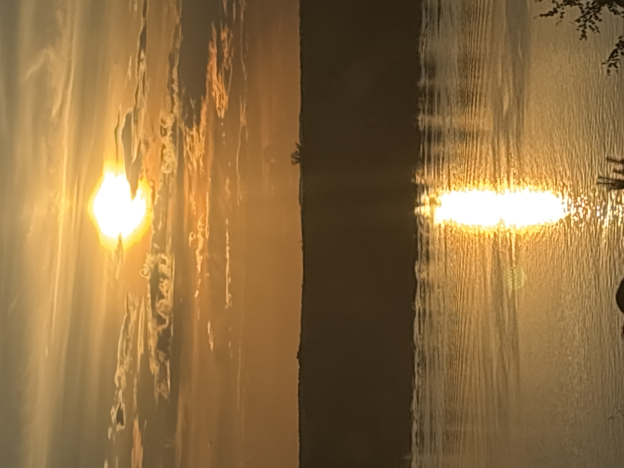This media file depicts a serene sunset over a body of water, likely a river or lake, in Olkhovka, Russia. The scene is dominated by the warm, golden hues of the setting sun, which casts a brilliant reflection on the rippling water. Wispy clouds, illuminated by the sunlight, add texture to the sky.
The image is split into two distinct sections by a dark, vertical element that appears to be the silhouette of a landmass, possibly the opposite bank of the water body. On the left side, the sun is prominently visible, radiating light and creating a soft glow. The sky here is filled with the soft tones of the sunset, with streaks of orange and yellow. On the right side, the reflection of the sun on the water is more pronounced, appearing as a bright, elongated band of light. The water itself shows a pattern of vertical ripples, suggesting a gentle current or breeze.
In the upper right corner, the dark silhouette of tree branches can be seen, adding a natural frame to the scene and suggesting the presence of vegetation along the shore. The overall mood of the image is tranquil and peaceful, evoking a sense of calm and natural beauty. The time of day is clearly sunset, and the weather appears to be clear with some cloud cover. There is no visible text in the media file.