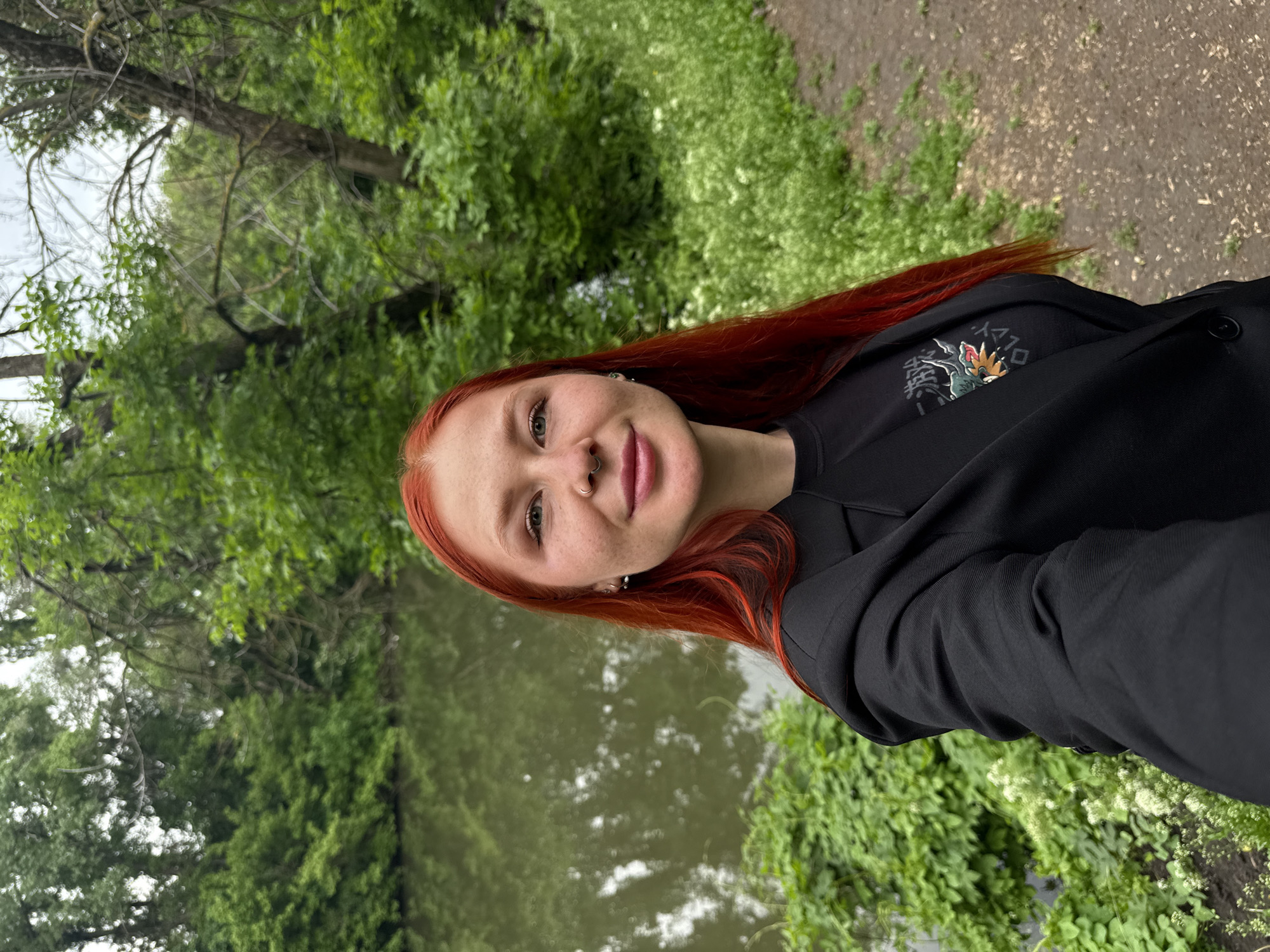 A young, fair-skinned woman with red hair is posing for a photograph outdoors. She is wearing a black jacket over a black t-shirt with a printed design. The background shows a body of water, possibly a river or lake, bordered by lush greenery. A path or ground with gravel can be seen on the right side of the image. The location may be a park or natural area in Rostov-on-Don, Russia, based on the typical landscaping of the region.