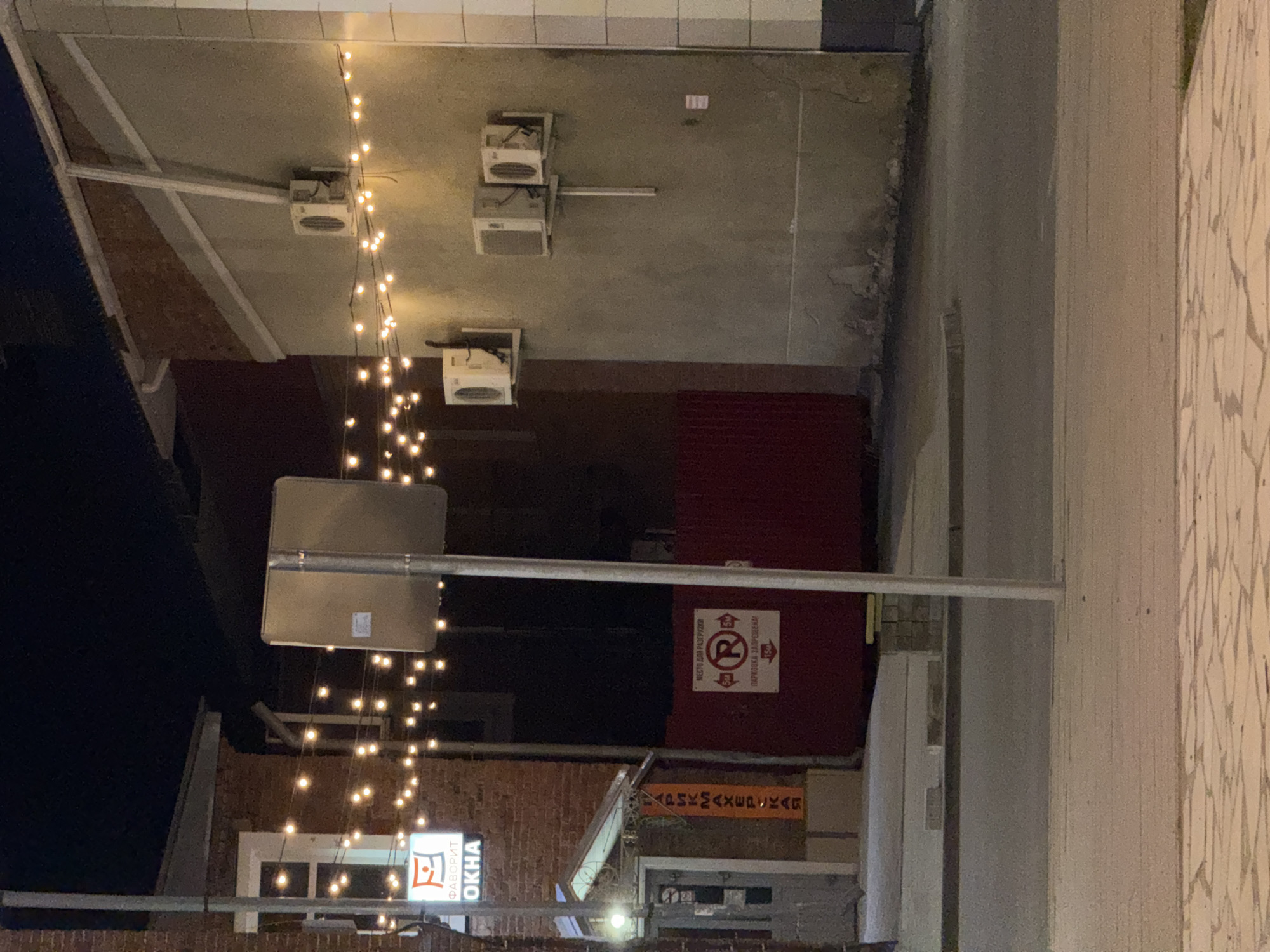 The image depicts a street scene at night in what appears to be Sarapul, Russia. A string of lights hangs diagonally across the upper left of the frame. Above, three air conditioning units are mounted on the ceiling of an overhang. A parking sign is visible, as well as signage in what appears to be the Russian language. A commercial sign for "Favorit OKHA" windows is identifiable. The environment appears to be urban, with brick and concrete architecture.