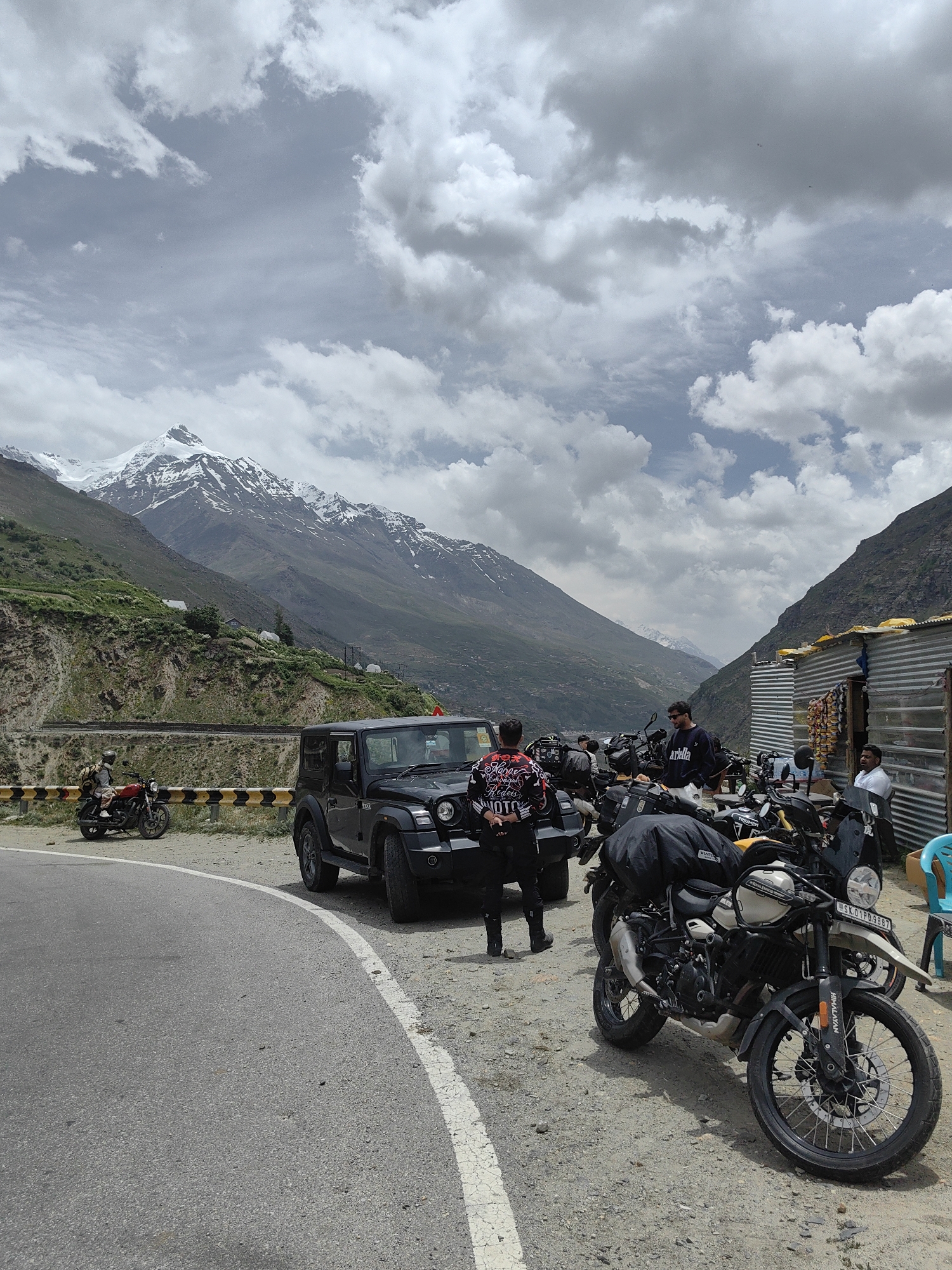 The image captures a group of motorcyclists and their bikes parked on the side of a road with a dramatic mountain backdrop. In the foreground, a black Mahindra Thar SUV is parked. Several motorcycles are lined up, some with luggage attached, suggesting a road trip. People are visible around the vehicles, some standing and others possibly seated in a small roadside structure.
The scene is set in a mountainous region, with towering peaks, some capped with snow, dominating the background. Green slopes and rocky outcrops form the terrain leading up to the mountains. The sky is partly cloudy, with a mix of white and grey clouds, suggesting a daytime scene. The road appears to be paved with a white line marking the edge.
Visible text includes a license plate on one of the motorcycles which reads "SK-01PD-9997". Another motorcycle has "HIMALAYAN" written on it. Text on the jackets of some individuals is also visible but difficult to decipher clearly. The overall mood appears to be one of adventure and camaraderie, common among motorcycle touring groups. The location, with its rugged mountains and roadside stops, is indicative of a scenic route, likely in a region known for its challenging terrain and natural beauty, consistent with the context of India.