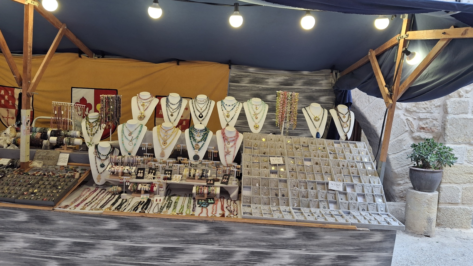 This image depicts an outdoor market stall displaying a variety of necklaces, bracelets, and other jewelry items. The stall is covered by a blue tarp and illuminated by several hanging light bulbs. Prominently featured are numerous necklaces arranged on white display busts, showcasing colorful beads and pendants. To the left, a rack of bracelets and a display of smaller jewelry pieces are visible. A large board filled with small, individually packaged necklaces or pendants is set up on the right side of the stall. In the background, a yellow banner with a crest-like design and red and white elements is hung. The stall is situated on a cobblestone surface, with a stone wall visible behind it to the right, where a potted plant sits on a stone pedestal. The overall scene suggests a craft market or artisan fair in a historic setting. The location is identified as La Fresneda, Spain, specifically within the province of Teruel in the autonomous community of Aragon.