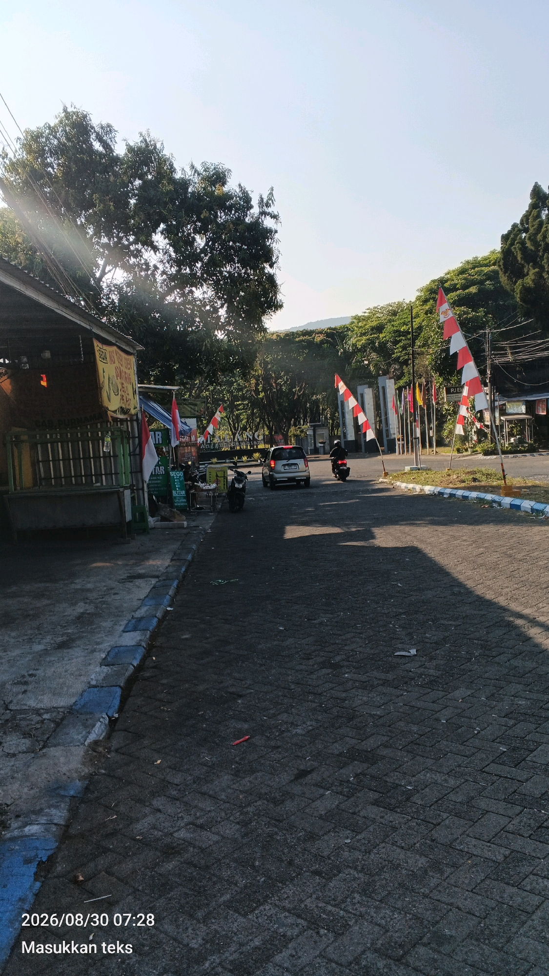 The image depicts a street scene at what appears to be the entrance to a facility or public space in Sumberporong, Indonesia. The time stamp on the image indicates August 30, 2026, at 07:28. The foreground is a paved road with a textured surface, bordered by a sidewalk that has blue and grey markings. To the left of the road, there is a small food stall or kiosk with a wooden structure and a striped awning. Various signs and merchandise are visible at the stall.
Several Indonesian flags, characterized by their red and white horizontal stripes, are prominently displayed on poles along the street, suggesting a national holiday or a patriotic event. In the background, there is a large gate or entrance structure, with several other flags of various designs on poles. Trees and lush green foliage line the sides of the road and the background, indicating a relatively green environment.
There are a few vehicles present: a silver car is driving away from the camera, and a motorcycle is also seen moving in the same direction. Another parked motorcycle is visible near the food stall. The sky is bright and clear, with the sun casting long shadows, suggesting it is early morning. The overall impression is a typical street scene in Indonesia, with a mix of commercial activity, public infrastructure, and a strong presence of national symbols.