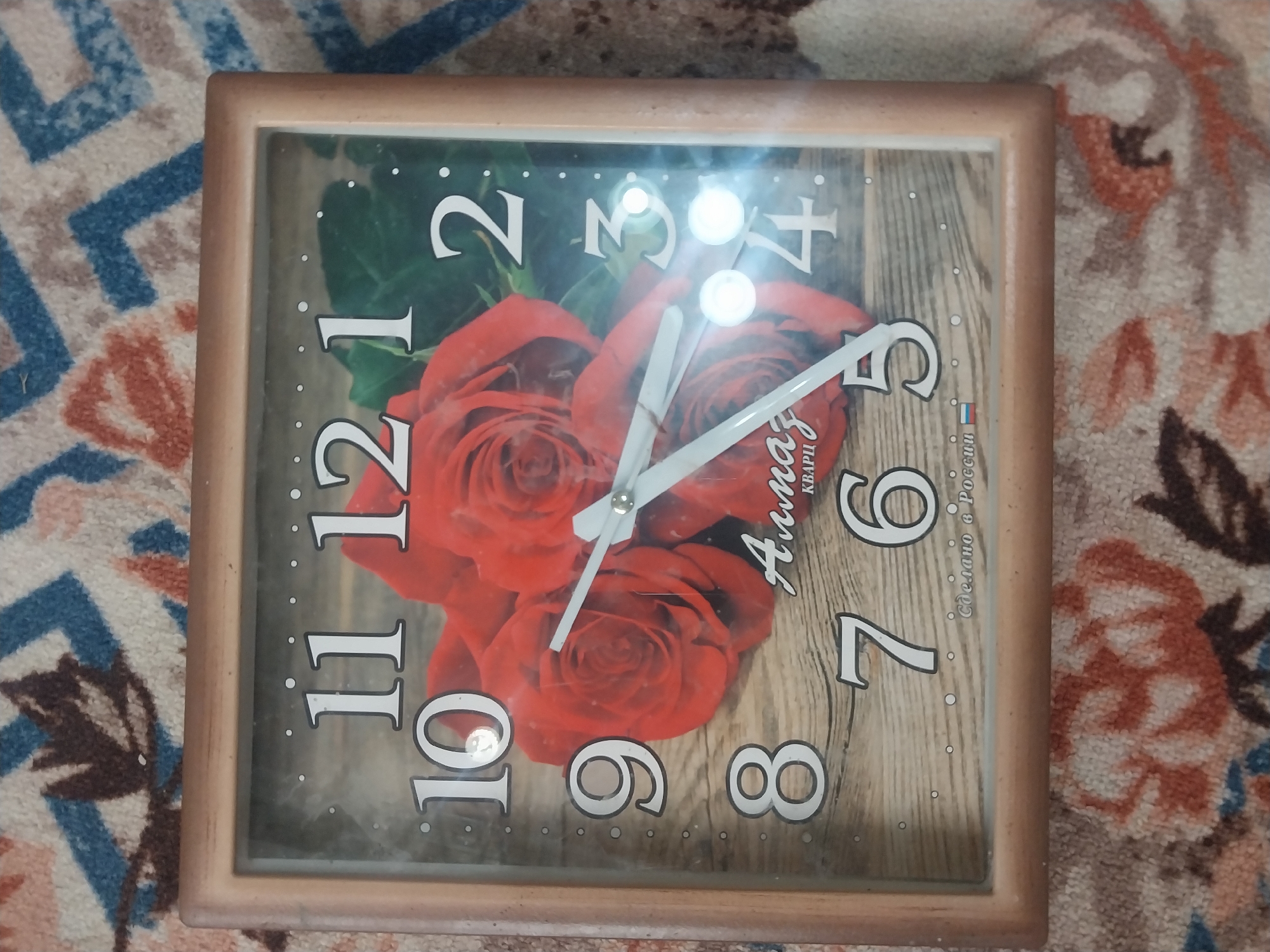 A square clock with a light brown frame is displayed on a carpet with a floral pattern. The clock face features a print of red roses against a wooden background. The numbers on the clock are white and stylized, indicating the hours. The clock has two white hands. In the center of the clock face, there is text that reads "Алмаз КВАРИ" and below it, "Сделано в России" with the Russian flag emoji. There are no people in the image, and no information about its location in Blagoveshenka, Kyrgyzstan is provided within the image itself.
