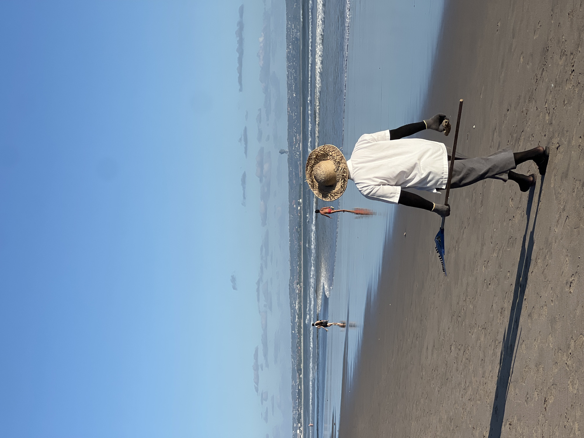 A person wearing a straw hat, white shirt, grey pants, black gloves, and sandals walks on a sandy beach in North Kuta, Indonesia. The person is holding a long stick with a blue object attached to one end and a brown object in their other hand. The beach is wet and reflects the sky, with gentle waves rolling onto the shore. In the background, there are other people walking on the beach and a distant cityscape with a plume of smoke rising from it. The sky is clear and blue with some scattered clouds.