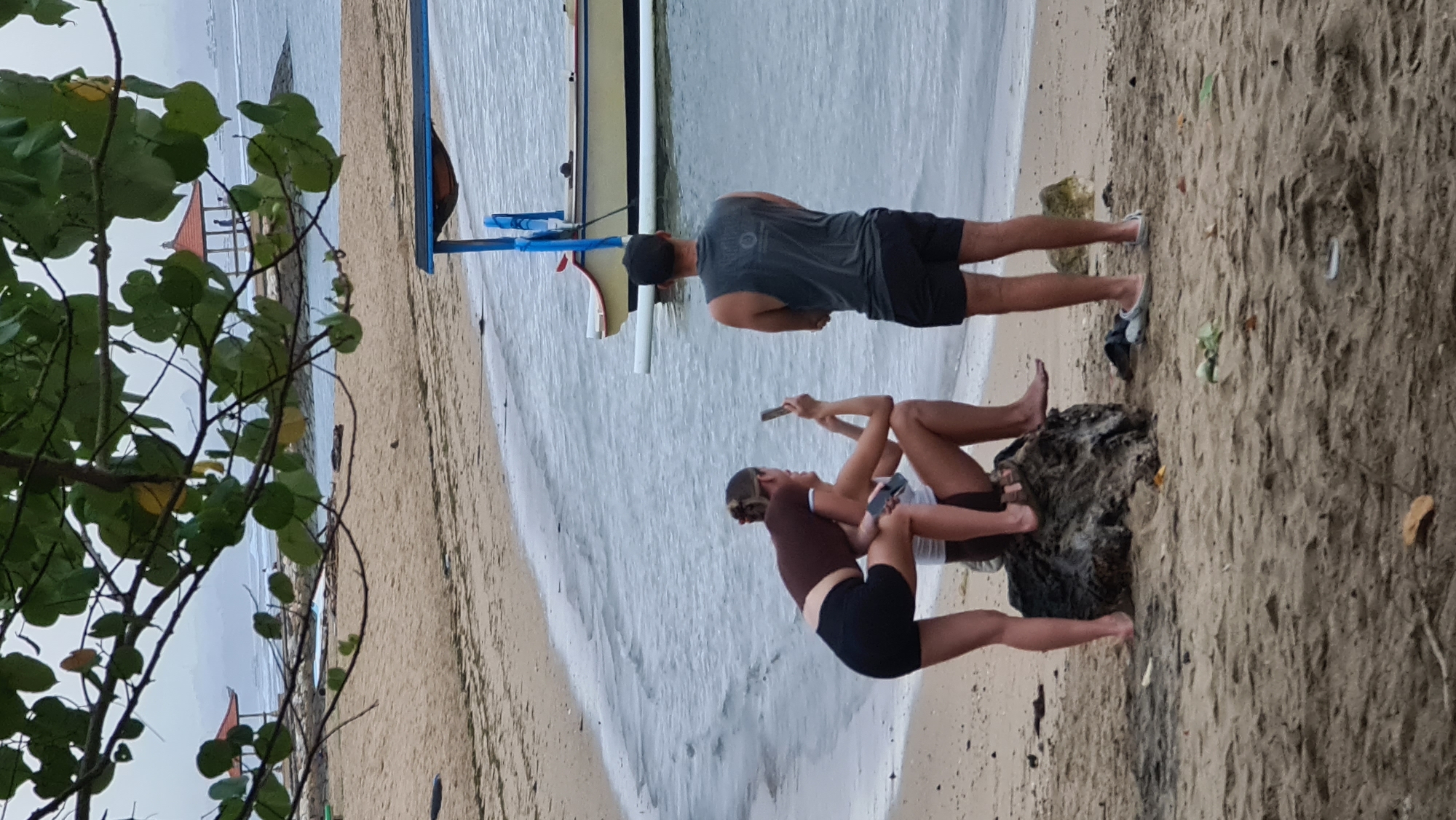 The image captures a scene on a sandy beach in Denpasar, Indonesia, likely near the coast. In the foreground, three individuals are present. Two young women are interacting on a large rock near the water. One woman, wearing a brown crop top and black shorts, is leaning forward, holding a phone and possibly taking a picture or recording. The other woman, wearing a white top and dark shorts, is seated on the rock and looking towards the first woman. A man wearing a dark grey sleeveless shirt and black shorts stands behind them, facing the ocean with his arms crossed.
The environment consists of a sandy beach, with the ocean waves gently lapping at the shore. The water appears calm and a light blue-grey. In the background, a traditional Balinese boat with outriggers is moored near the shore. Further in the distance, a small red-roofed pavilion structure is visible on a rocky outcrop, and several other boats are scattered across the water. Overcast skies suggest a cloudy day. The vegetation, including tree branches with green leaves, frames the top of the image. The overall impression is a relaxed, casual gathering on a tropical beach.