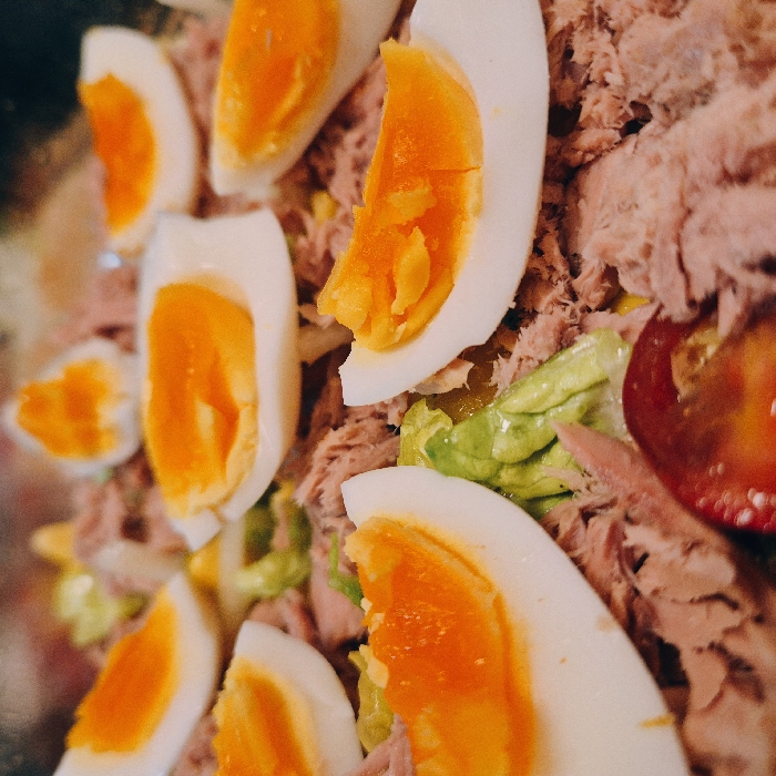 The image displays a close-up, top-down view of a prepared meal. The primary components visible are multiple quartered hard-boiled eggs, presenting white albumen and firm orange-yellow yolks, distributed across a bed of flaked light pink tuna. Interspersed among the tuna and eggs are pieces of green leafy lettuce. A single round slice of red tomato is partially visible in the lower foreground. The focus is entirely on the food, with no discernible background elements. The scene provides no visual information to determine a specific location within the city of Strasbourg, France.