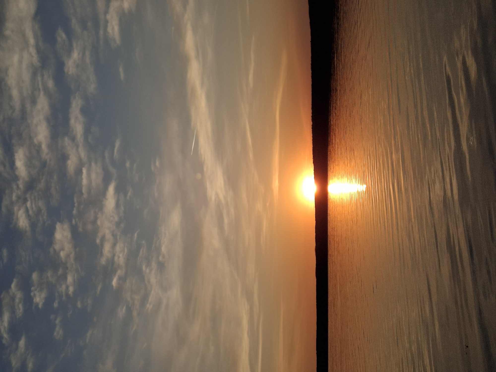 The image depicts a sunset over water, likely a lake or river. The sun is positioned on the horizon, partially obscured by a dark landmass, and its reflection forms a bright line extending across the water towards the viewer. The sky is filled with clouds, varying in color from pale blue to orange, reflecting the sunset. A contrail from an airplane is visible. The location is Chaikovsky, Russia. No people are visible.