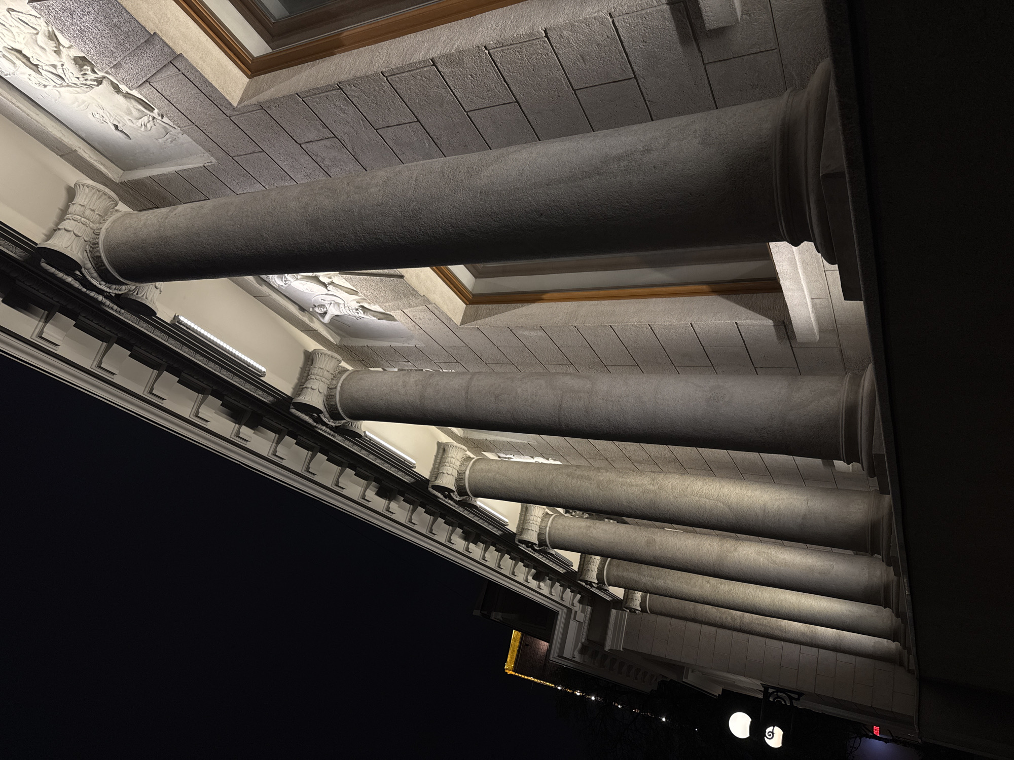 The image presents a low-angle view of a colonnade, likely part of a building facade, taken at night. The architectural style incorporates classical elements, including substantial cylindrical columns with ornate capitals and decorative friezes. The columns are uniformly gray and appear to be made of stone or concrete. Above the columns, a ceiling features a textured, block-like pattern, possibly mimicking stone construction. Recessed panels in the ceiling contain decorative reliefs. The image exhibits strong contrast with a dark, almost black background on the left side of the frame, and a bright, illuminated section to the right where lights and architectural details are visible in the distance. This architectural style is not unique to any single city and cannot confirm the location to be in Moscow, Russia.
