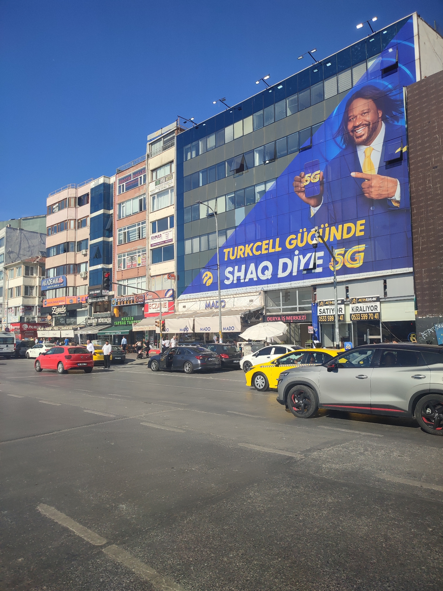 The image depicts a bustling street scene in Kadıköy, Turkey, under a clear blue sky. A prominent feature is a large blue billboard on the side of a modern building, advertising "TURKCELL GUCUNDE SHAQ DİYE 5G," with a smiling image of Shaquille O'Neal holding a 5G phone. Below the billboard, the building's facade displays various shop signs, including "MADO," "ECZANE," and "CALİFORNİAN BURGER."
The street is filled with traffic. Several cars are visible, including a red hatchback, a silver SUV with red accents on its wheels, and a yellow taxi. People are seen on the sidewalks and near the traffic lights, some appearing to be pedestrians waiting to cross, while others are likely patrons of the businesses. The street itself is asphalt with painted lane markings. Traffic lights are also present. The buildings lining the street are a mix of modern and older architecture, with multiple stories and many windows. The overall impression is of an active urban environment.