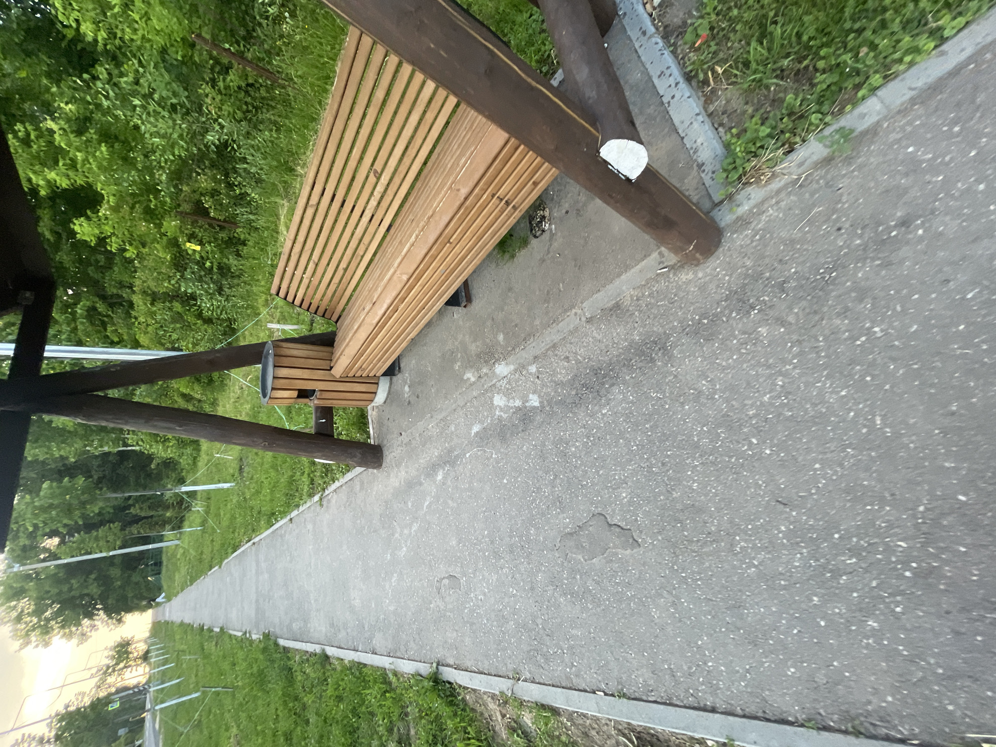 The image depicts a paved walkway in Красные Баки, Russia, with a wooden swing set and bench on the right side. The walkway extends into the distance, bordered by green grass and trees. The swing set has a wooden bench with horizontal slats and a visible wooden bin next to it. The ground is asphalt with some visible patches and cracks. The environment appears to be a park or recreational area. The lighting suggests it is either early morning or late afternoon. There are no people or animals visible in the frame.